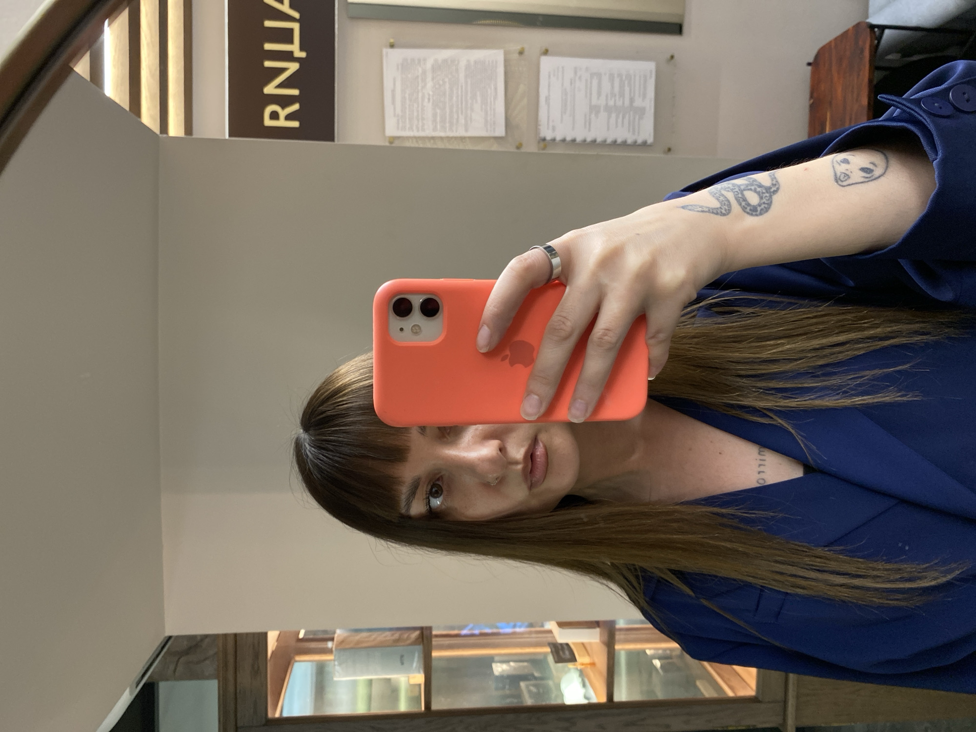 A medium shot depicts a person with shoulder-length brown hair and bangs, taking a selfie in a mirror. The person is holding a coral-colored iPhone, obscuring the center of their face. They are wearing a blue blazer. A silver ring is visible on their finger. The person has tattoos on their arm, including a snake and an alien-like figure. A tattoo is also visible on the chest, reading "WILLOW."
The mirror reflects the environment, including a window showing an interior space with shelves and what appear to be display cases. Behind the person in the reflection, there are wall-mounted signs, some with text.
The location is Votkinsk, Russia.