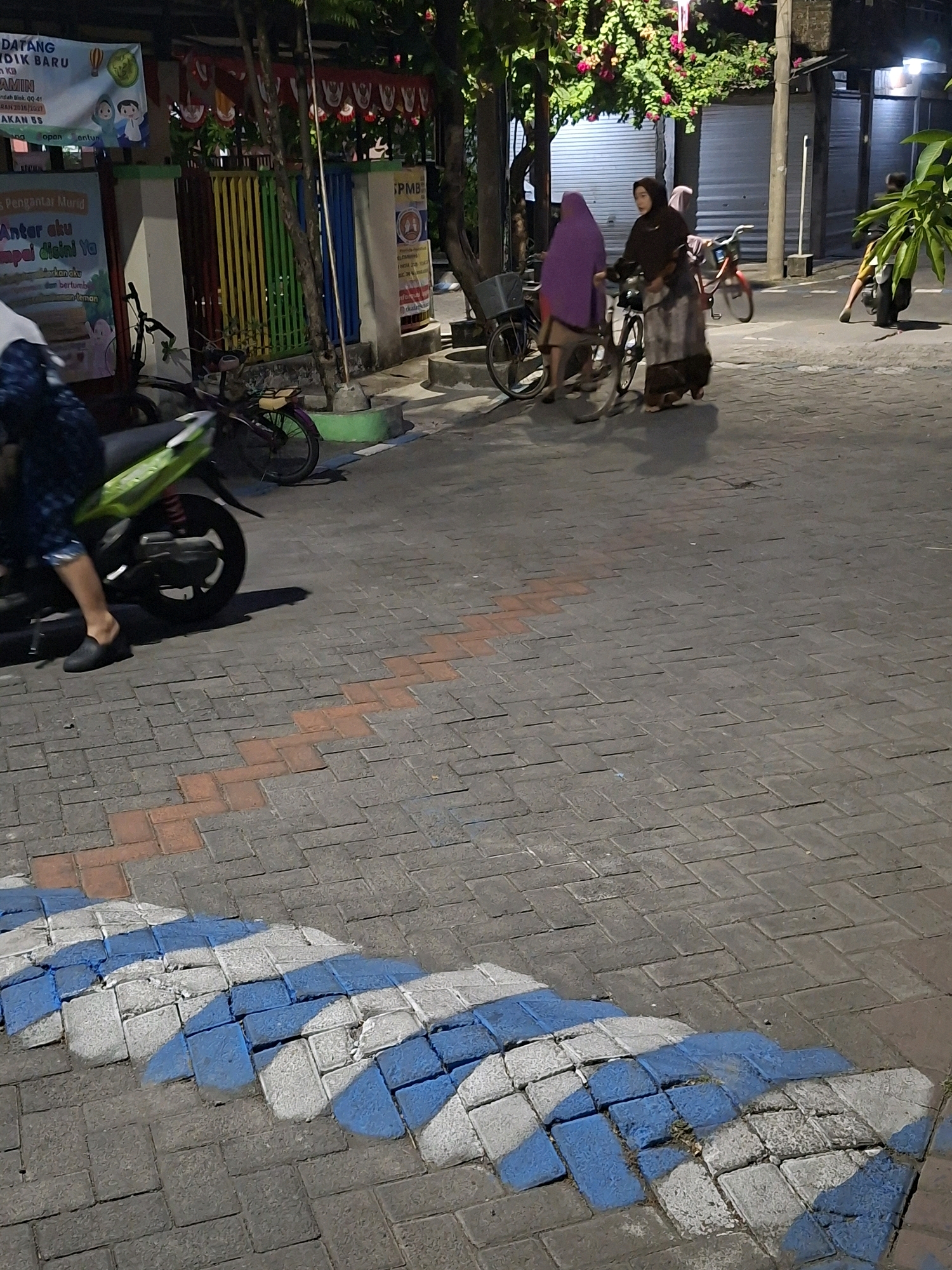 The image shows a street scene in Surabaya, Indonesia, at night. In the foreground, a patterned walkway with blue and white markings is visible on a paved road. Further back, a person is riding a green motorcycle. Two women are walking with bicycles, and another person is on a bicycle in the background. Buildings line the street, and trees with pink flowers are present. Signs in Indonesian are visible on the buildings. The overall atmosphere is that of a typical street in Surabaya after dark.