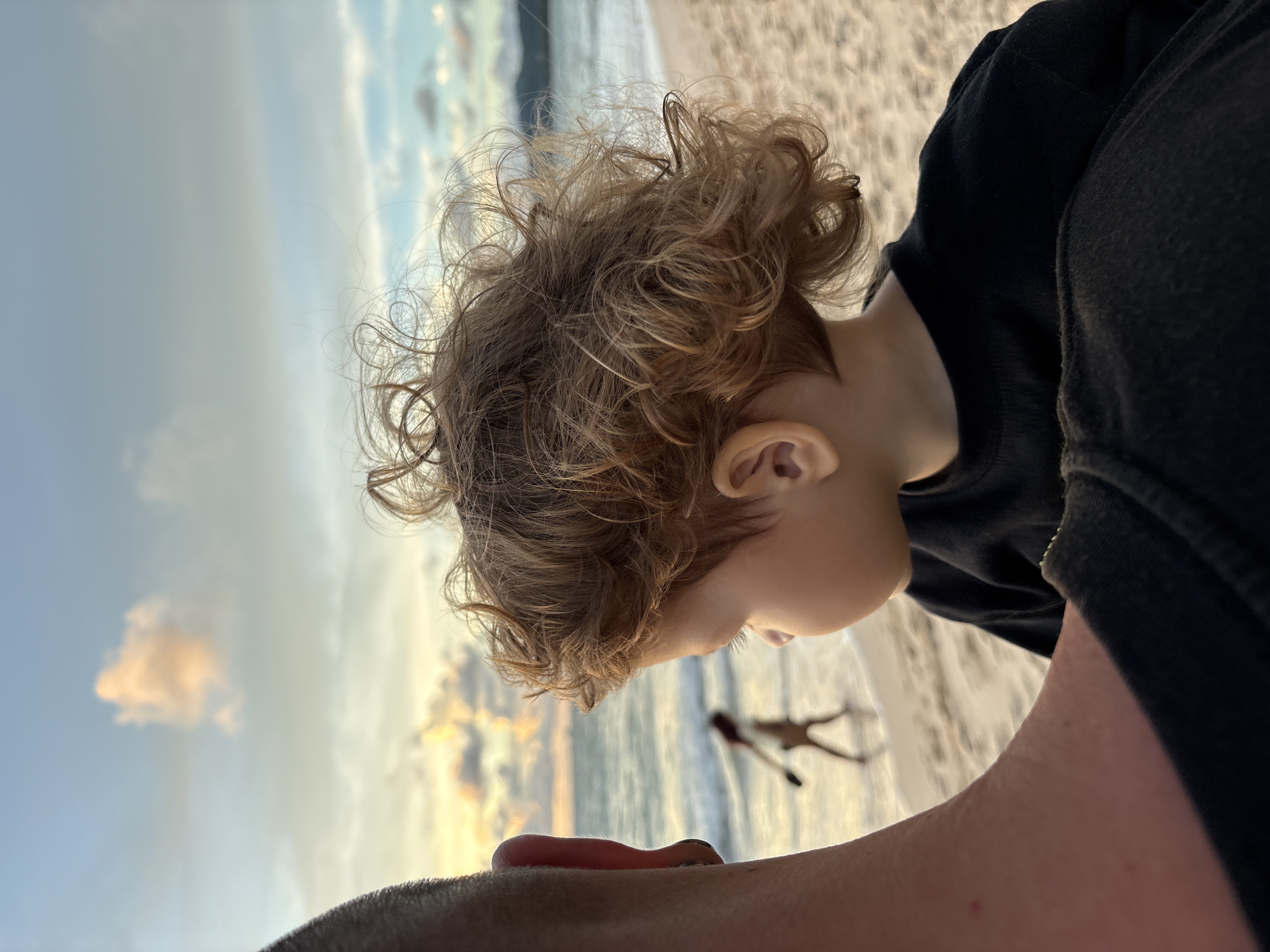This is a close-up, rotated shot of a young child with curly blonde hair, seen in profile. The child is wearing a black top and is looking down towards the sand. In the background, out of focus, is a beach scene with the ocean and a cloudy sky. The sun appears to be setting, casting a warm glow on the clouds and the water. A faint silhouette of a person can be seen walking along the shoreline in the distance. The foreground features a person's arm and shoulder, also wearing a black garment, suggesting a selfie-style shot from an adult holding the child. The overall mood is relaxed and summery, likely taken during a vacation on a beach. The location is identified as Choeng Thale, Thailand. There is no visible text in the image.