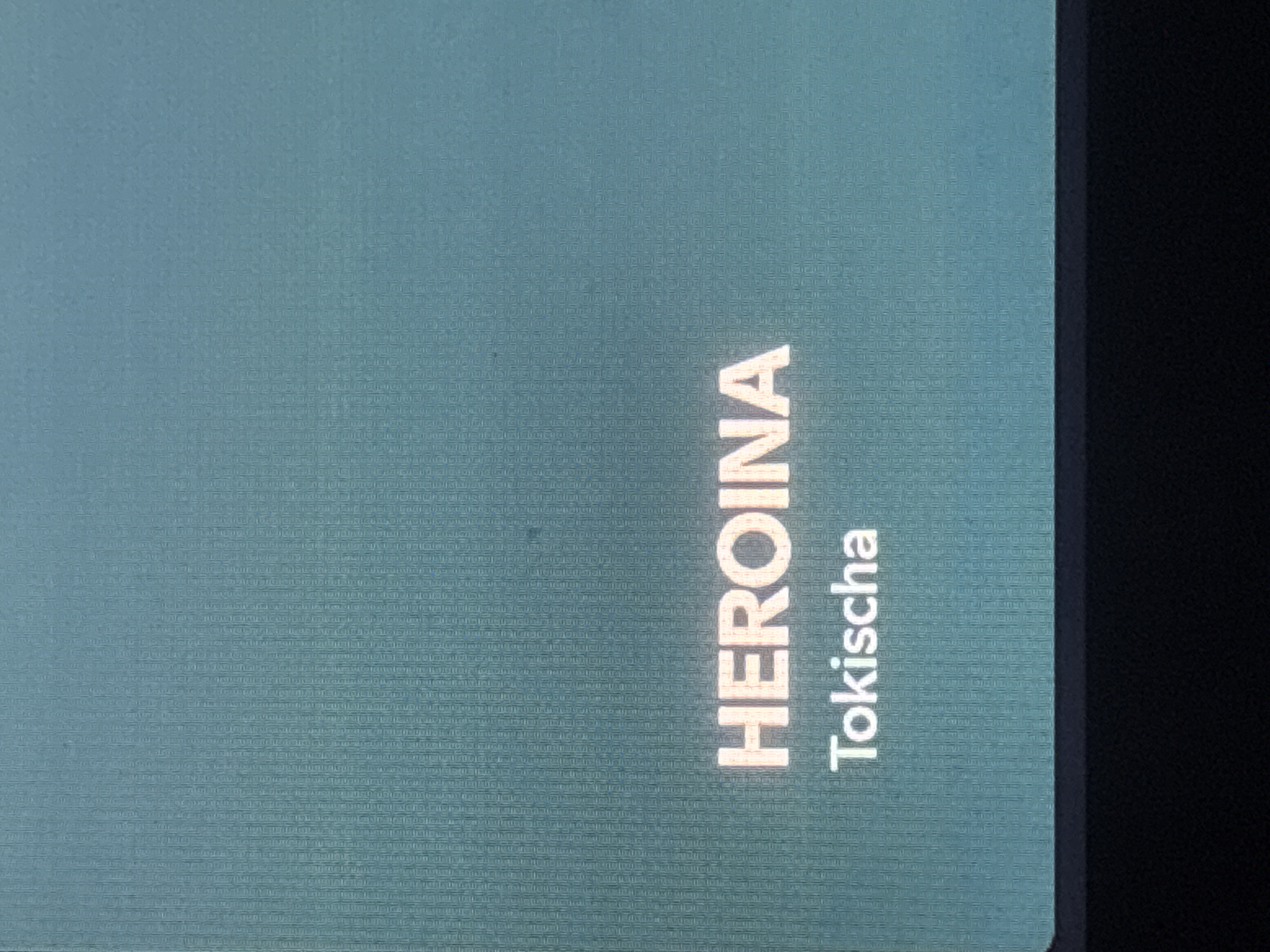 The image shows a close-up of a screen displaying the vertically oriented word "HEROINA" in a light color, with "Tokischa" written below it in a slightly darker color. The screen appears to be part of a larger device or surface, as evidenced by the presence of a dark edge on the right side of the frame. The background of the screen is a textured teal color. The location is not discernible from the image.