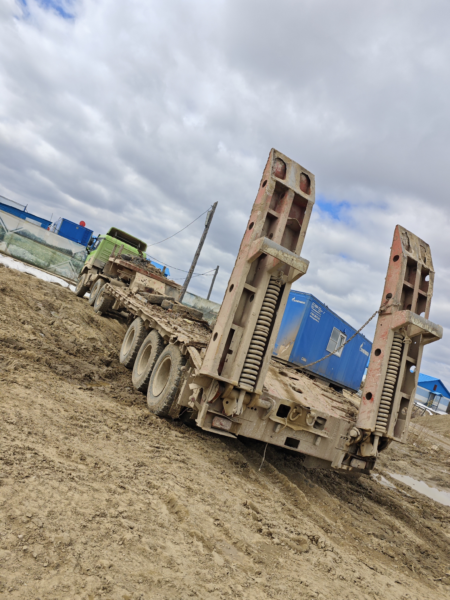 The image depicts a heavy-duty, lowboy trailer stuck in muddy terrain in an outdoor location with an overcast sky. The trailer is loaded with a blue container, labeled "Gazpromtrans." The trailer is attached to a green cab-over semi-truck. The environment suggests a construction or industrial site with other buildings or structures visible in the background. Based on the inscription on the blue container, the location is likely in Russia, but the specific city is not discernible from the image.