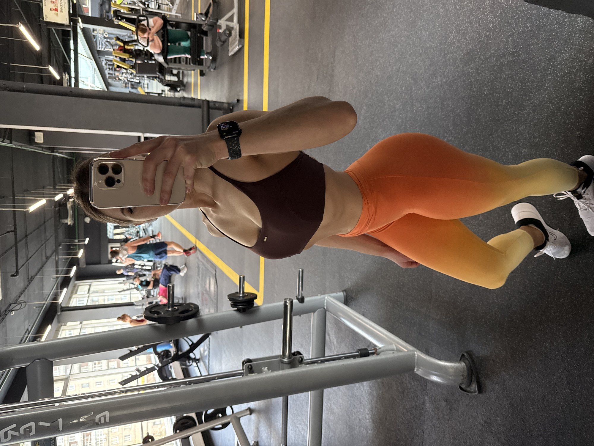 A woman in athletic wear takes a selfie in a gym. She is wearing a brown sports bra, orange and yellow ombre leggings, and white sneakers. Her phone is visible, and she is wearing a smartwatch. In the background, other gym-goers are using various exercise machines. The gym appears to be located in Saint Petersburg, Russia, based on the view of buildings visible through the windows.