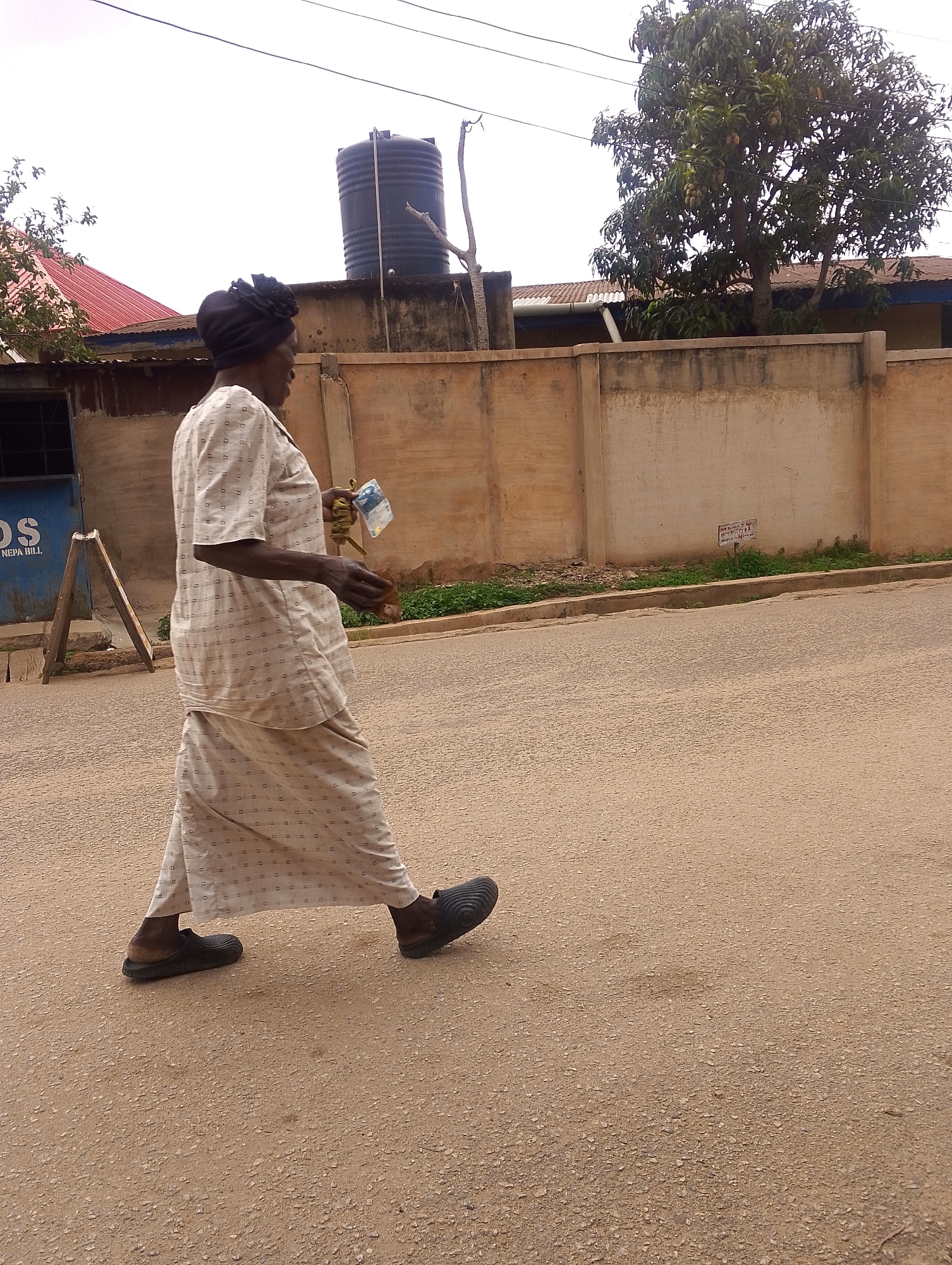 The image shows an older Black woman walking on a paved street in Unknown, Unknown. She is dressed in a light-colored, patterned matching top and long skirt. She has a dark head covering and wears dark shoes. In her hand, she is holding an item. The background includes a tall water tank behind a concrete wall, a tree, and parts of buildings. A sign is partially visible on a nearby structure. The street appears to be dusty or unpaved.