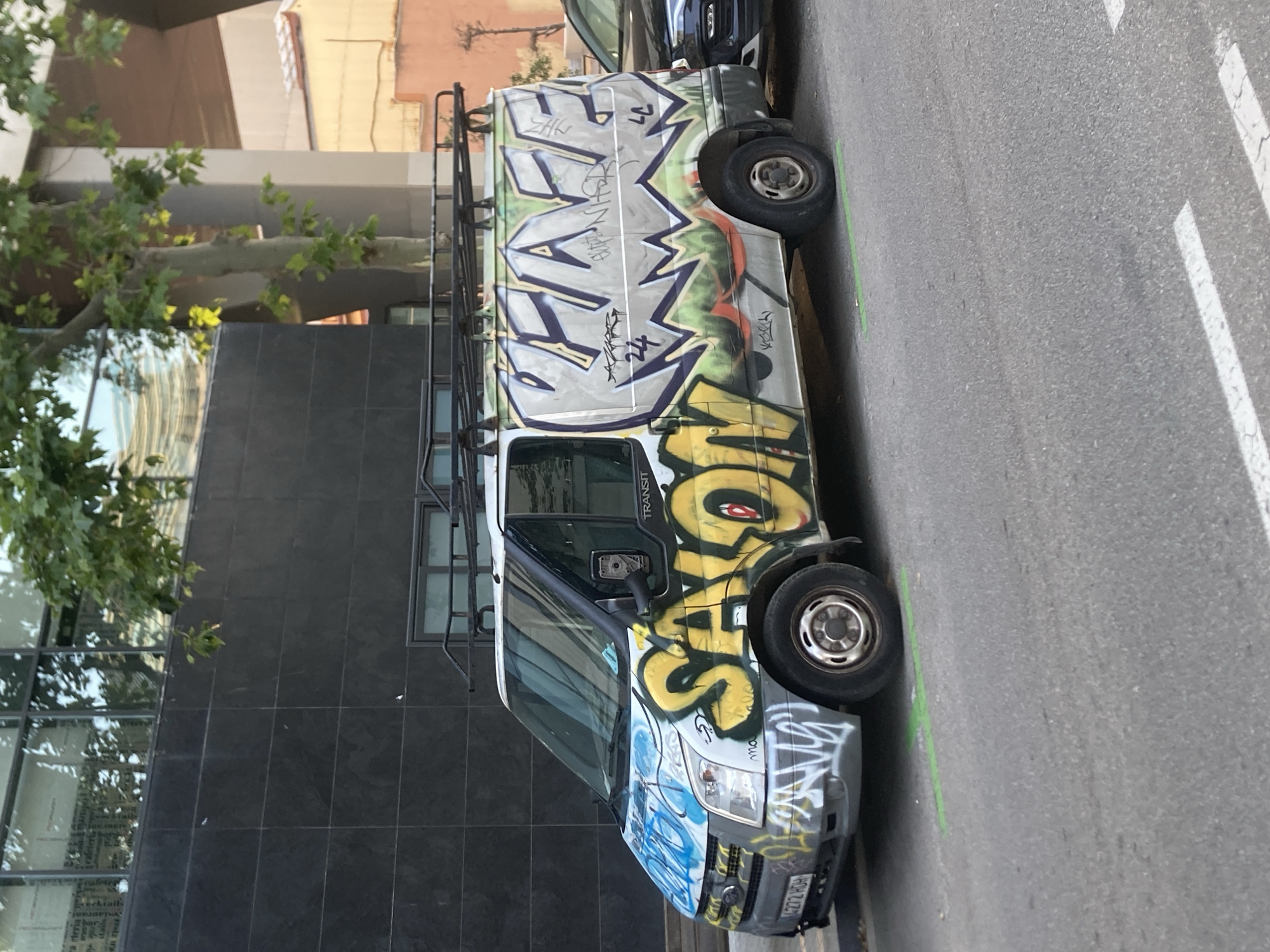 A white Ford Transit van, extensively covered in graffiti, is parked on a city street in Barcelona, Spain. The graffiti features bold, colorful lettering and designs. On the front of the van, the word "SAXON" is prominently displayed in large yellow bubble letters with red and black accents. The hood and front bumper are covered in blue and white spray paint. The rear side of the van displays the word "CHASE" in large, stylized grey letters with purple outlines and green and orange accents. The van has a roof rack. The street is paved with asphalt, and a single white dashed line runs across it. A green line is painted on the asphalt in front of the van's tires. In the background, a modern building with dark tiled walls and large windows is visible, along with trees and other urban elements. A dark car is parked to the right of the van, partially obscuring it. The scene appears to be during daylight.