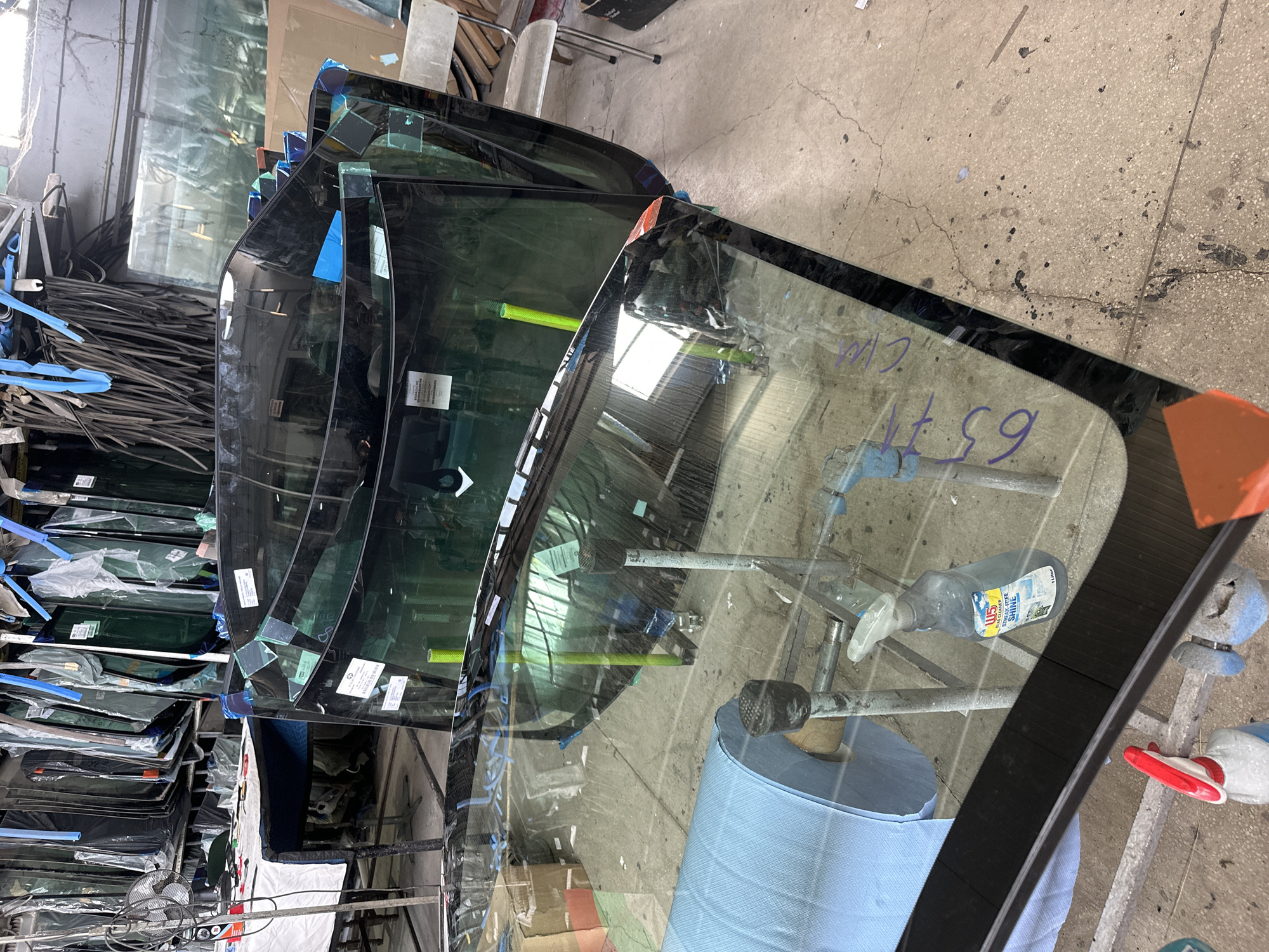 The image displays an indoor scene, likely within an automotive glass workshop or warehouse. The primary subjects are numerous car windshields and other pieces of automotive glass, arranged in stacks and held upright by metal support frames. Many of these glass panels are protected by blue tape on their edges and corners, and some bear white informational labels.
In the foreground, through the clear glass of one windshield, a roll of blue protective material and a grey spray bottle are visible. The spray bottle has "W5" and "GLASS CLEANER SHINE" printed on its label, indicating its use. Above the numbers "6377" handwritten in purple marker, the letters "WCM" are also visible on this windshield. A green tubular object is also seen through the glass.
The floor is a cracked and stained concrete surface, typical of an industrial setting. In the background, further stacks of glass and general workshop items, including a small fan, are visible, suggesting a cluttered yet functional environment. No people are present, and no specific activity is taking place, depicting a moment of storage or preparation in an automotive glass business.