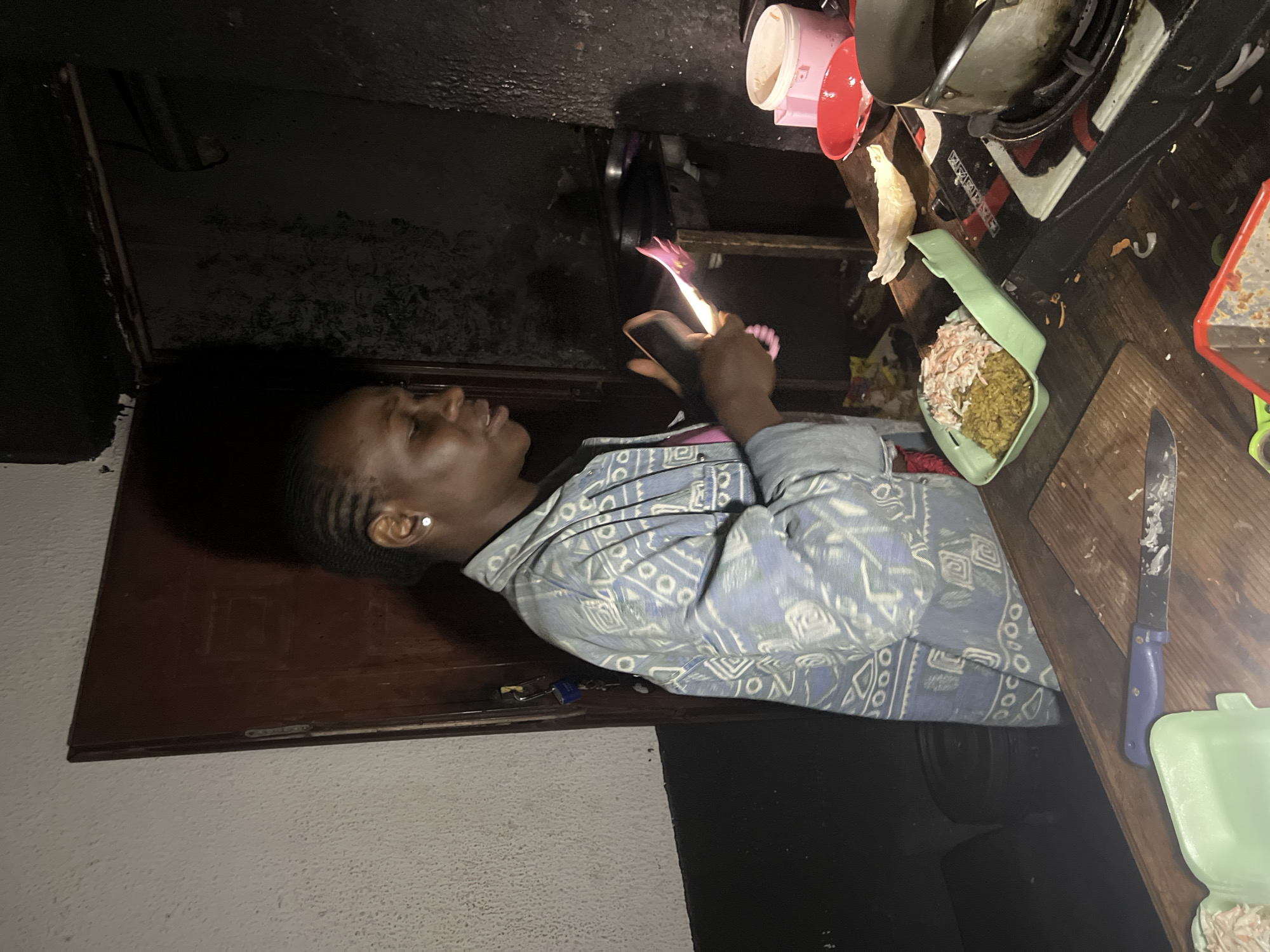 A young woman with braided hair is standing in a dimly lit kitchen, holding a lighter to a dish of food. She is wearing a blue and white patterned shirt. The kitchen appears to be in Durumi, Nigeria. On the counter, there are various containers of food, a knife, and a pot on a stove. The walls are textured and dark, suggesting the kitchen is small and perhaps not well-maintained.
