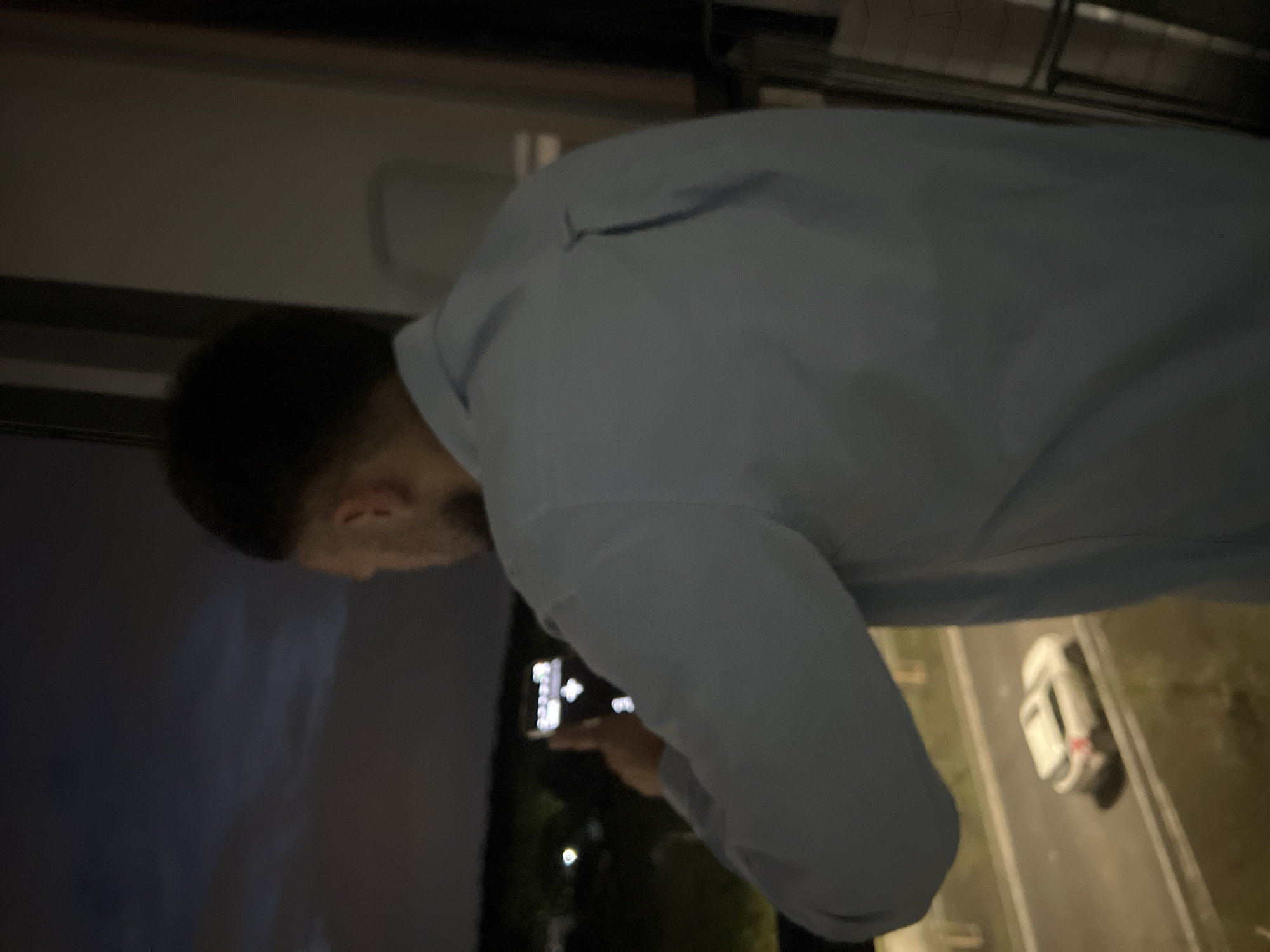 A man is seen from behind, wearing a light blue collared shirt, holding a smartphone up as if taking a picture. He is standing near a window with a view of a highway at dusk or night. On the highway, a white SUV is visible. The background shows a dark sky with some clouds and distant lights, possibly indicating a city environment. The overall lighting suggests it is either early morning or late evening.