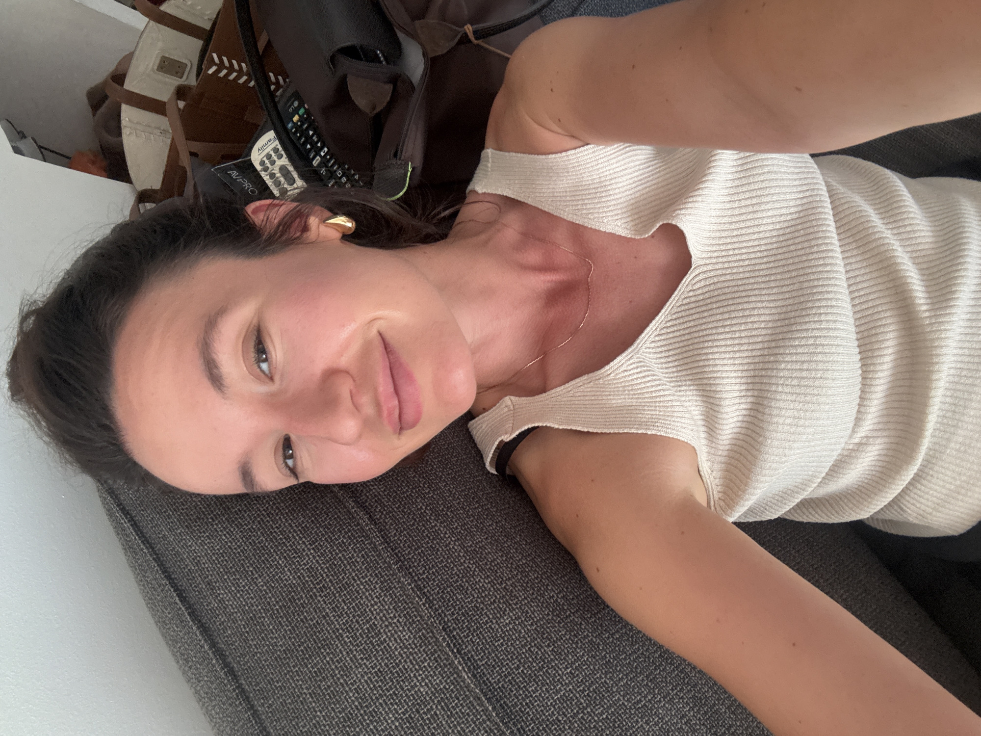 A young woman with her hair pulled up in a bun is lying on a grey couch, looking at the camera. She is wearing a white ribbed tank top and a thin gold necklace. She has gold teardrop earrings. In the background, there are several bags, a remote control, and what appears to be a black leather bag. The scene is indoors, likely in a residential setting.
