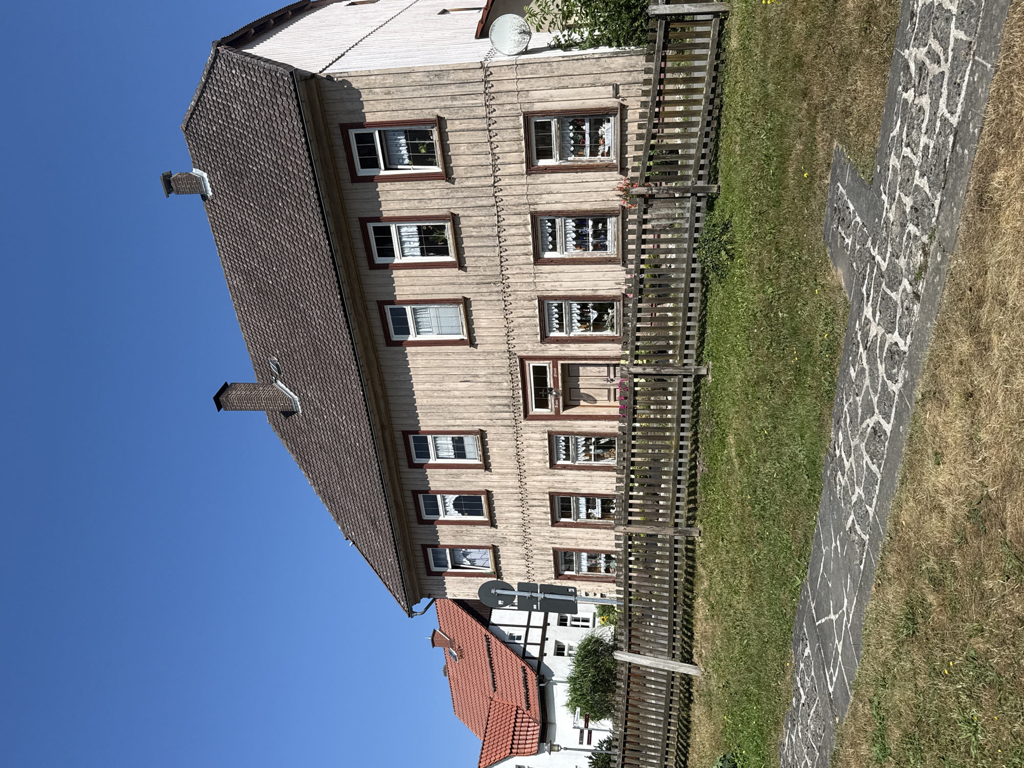 The image captures a sunny day in Walkenried, Germany, focusing on a two-story wooden house with a pitched, tiled roof and a brick chimney. The house is weathered, with vertical wooden planks forming its exterior. It features numerous windows with white curtains and dark red trim. A wooden fence runs along the front of the property, separating it from a grassy area. To the left, a portion of another building with a red-tiled roof and white and black half-timbered details is visible. The sky is clear and blue.
