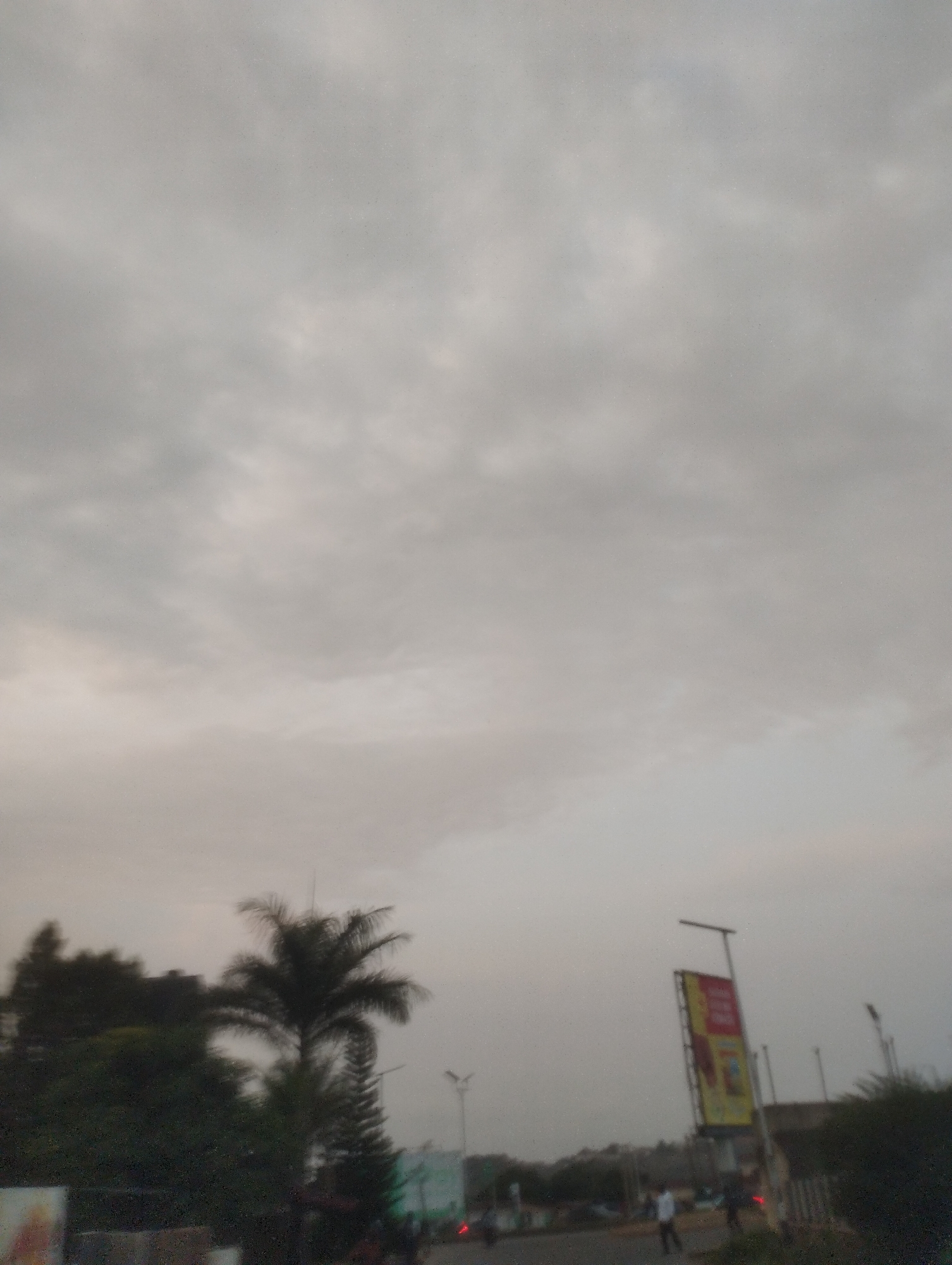 This is a blurry, overcast, outdoor, daytime photograph taken in Jos, Nigeria. The scene depicts a road with some trees, including a prominent palm tree, and a tall, vertical billboard on the right side. The billboard has a yellow and red design with some text that is unreadable due to the blur. There are a few indistinct figures of people and some vehicles present, suggesting a somewhat active urban or suburban environment. The sky is filled with heavy, gray clouds.