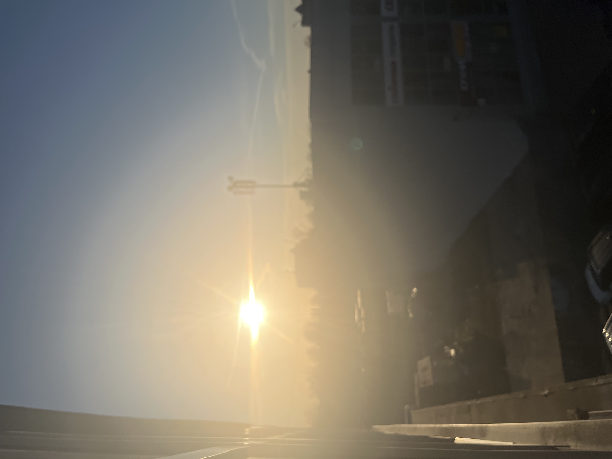 The image depicts an outdoor urban scene in Klin, Russia, under conditions of bright sunlight and atmospheric haze. The viewpoint is elevated, suggesting a window or balcony perspective.
The sun is prominently positioned in the upper-left quadrant, emitting a strong lens flare and causing significant overexposure and a warm, yellowish glow across much of the scene. The sky transitions from this bright yellow at the horizon to a pale blue in the upper-right. Visible in the upper-right sky are faint contrails.
On the far left, a vertical architectural element, possibly a window frame or balcony railing, is partially visible and out of focus. In the midground, a line of indistinct trees or vegetation is silhouetted against the bright haze. To the right of these, a tall cellular communication tower with multiple antennae stands against the sky. Further to the right, the upper sections of a multi-story building are visible, featuring a flat roofline. Signs with Cyrillic script are faintly discernible on the building's facade; one appears to read "КУХНИ" (KITCHENS).
The lower portion of the image is significantly darker and more blurred, revealing indistinct shapes consistent with parked vehicles on a ground surface. No identifiable individuals or specific actions are visible within the frame.