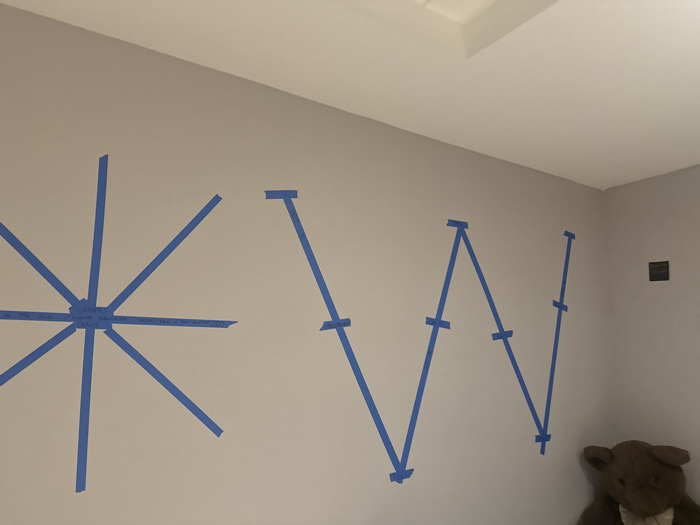 A light gray wall displays blue painter's tape outlining what appears to be the letters "KW" stylized. The "K" is constructed with multiple lines emanating from a central point, creating a starburst effect. The "W" is formed by angular lines mimicking the letter's shape. Small pieces of tape are placed perpendicularly across the lines. In the room's corner, a dark outlet and a stuffed brown bear are partially visible. The upper right shows a portion of the white ceiling.