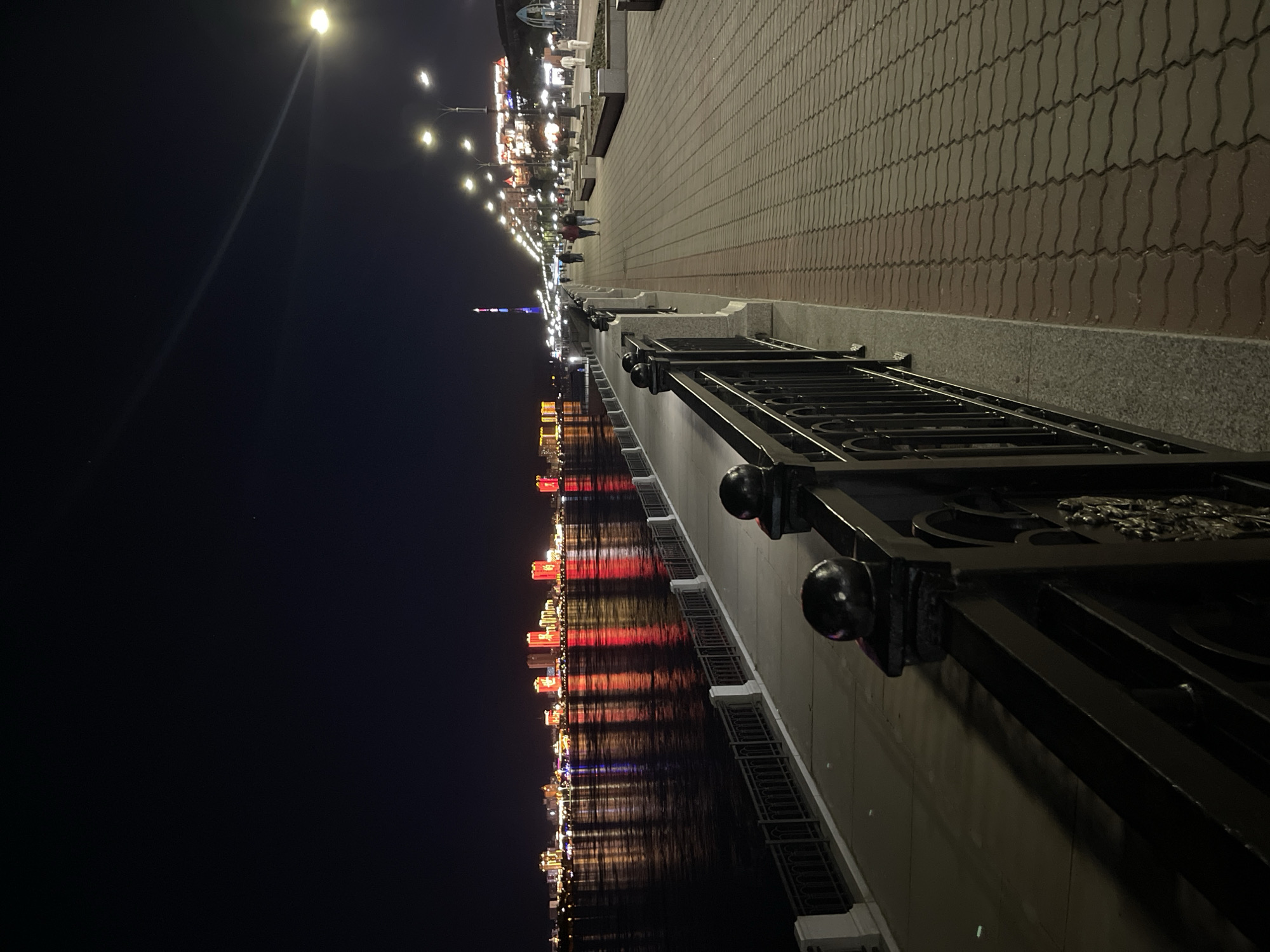An exterior night shot captures a view of the Amur River embankment in Blagoveshchensk, Russia. The embankment is paved with red and grey interlocking pavers, bordered by a black metal railing with rounded black finials. A stone retaining wall runs along the river side, also with a black metal railing. The Amur River reflects the lights of buildings on the opposite bank, which appear as a cityscape with brightly lit structures in red and yellow. Streetlights illuminate the pathway. Several people are visible walking along the embankment. The sky is dark with a few distant lights.