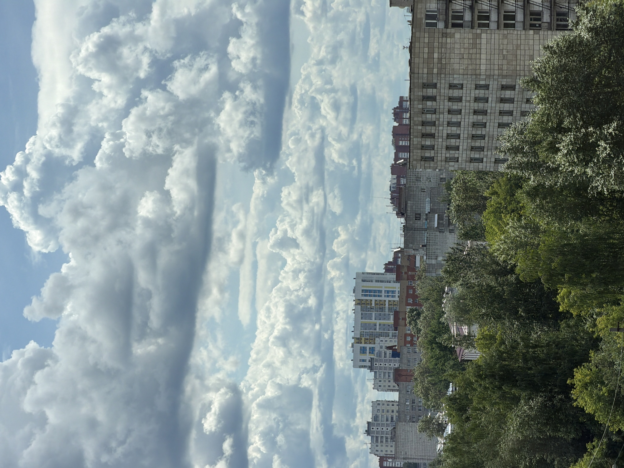 This is a vertical, eye-level, medium shot of a cityscape in Perm, Russia. The image is dominated by a dramatic sky filled with voluminous, white cumulus clouds, stretching across the left side of the frame. The clouds are a mix of bright white where the sun hits them and darker grays in their shadowed areas, creating a dynamic, textured sky.
On the right side of the image, a dense line of trees separates the sky from a row of apartment buildings. The trees are a lush, deep green, suggesting a warm season like summer. The buildings are of varying heights and architectural styles, with some appearing older and clad in light-colored tiles, while others are more modern with colorful facades, balconies, and many windows. The cityscape is dense, with buildings packed closely together.
The overall impression is one of a somewhat overcast but bright day, possibly late morning or early afternoon, given the strong illumination of the clouds. The composition creates a contrast between the natural beauty of the sky and clouds and the man-made structures of the city. There are no people or specific activities visible, placing the focus on the atmospheric conditions and the urban landscape. The mood is somewhat dramatic due to the intense clouds, yet also peaceful, with the greenery offering a sense of nature within the urban environment. No visible text is present in the image.