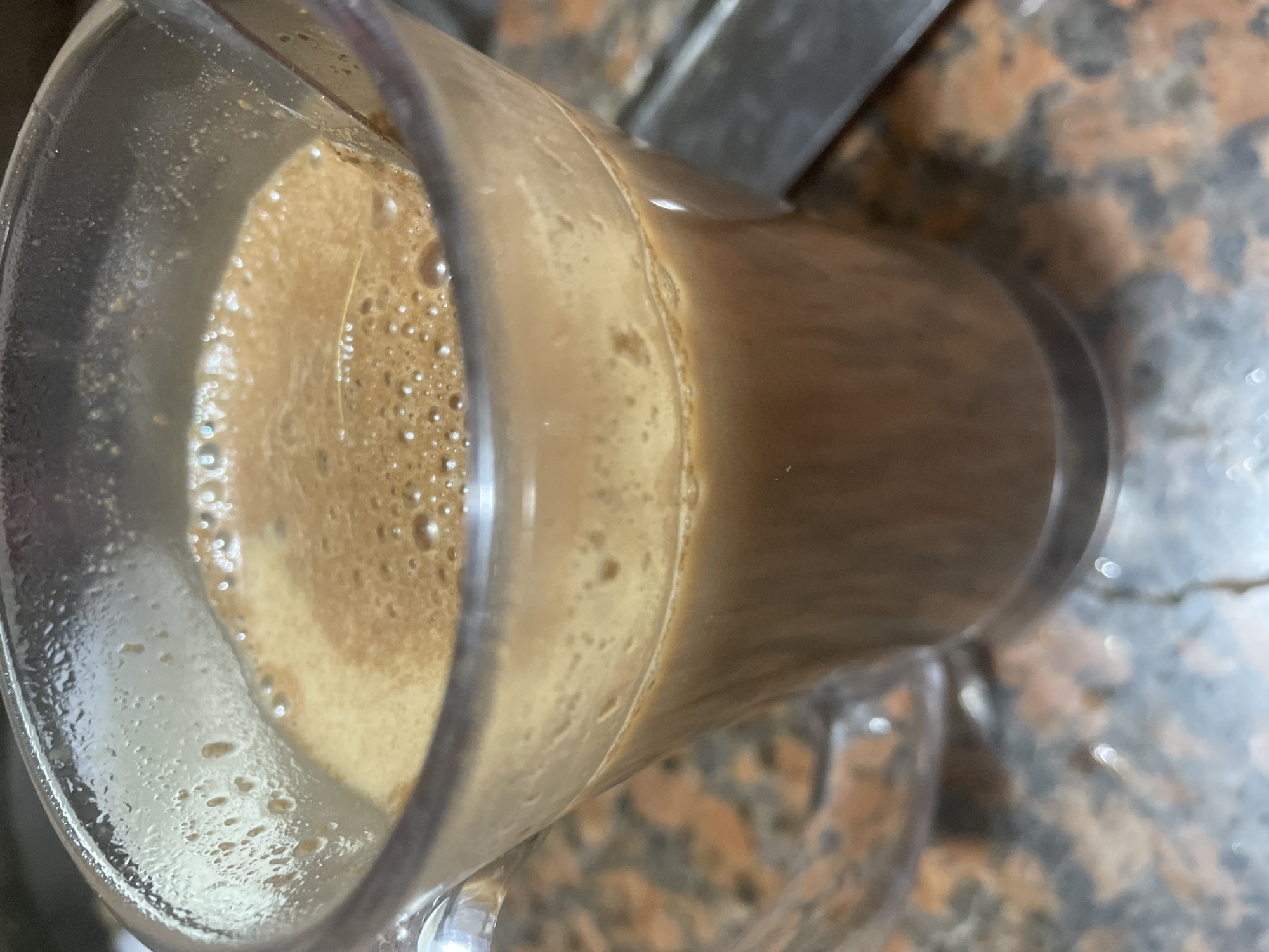 A close-up, slightly angled, top-down shot presents a clear glass mug containing a frothy, brown beverage. The drink exhibits a thick layer of light brown foam, densely populated with small bubbles, while the liquid beneath is a darker, rich brown. The transparent nature of the glass mug allows the beverage's color to be observed through its sides. The mug is positioned on a speckled surface, likely a countertop, featuring an irregular pattern of various shades of brown, black, and hints of lighter coloration. Portions of other objects or surfaces are faintly visible in the background, suggesting an indoor setting such as a kitchen or dining area. No individuals or visible text are present, and no specific activity is taking place beyond the static presence of the prepared drink. The image provides no specific visual cues for its location, but it is situated in Davao City, Philippines.