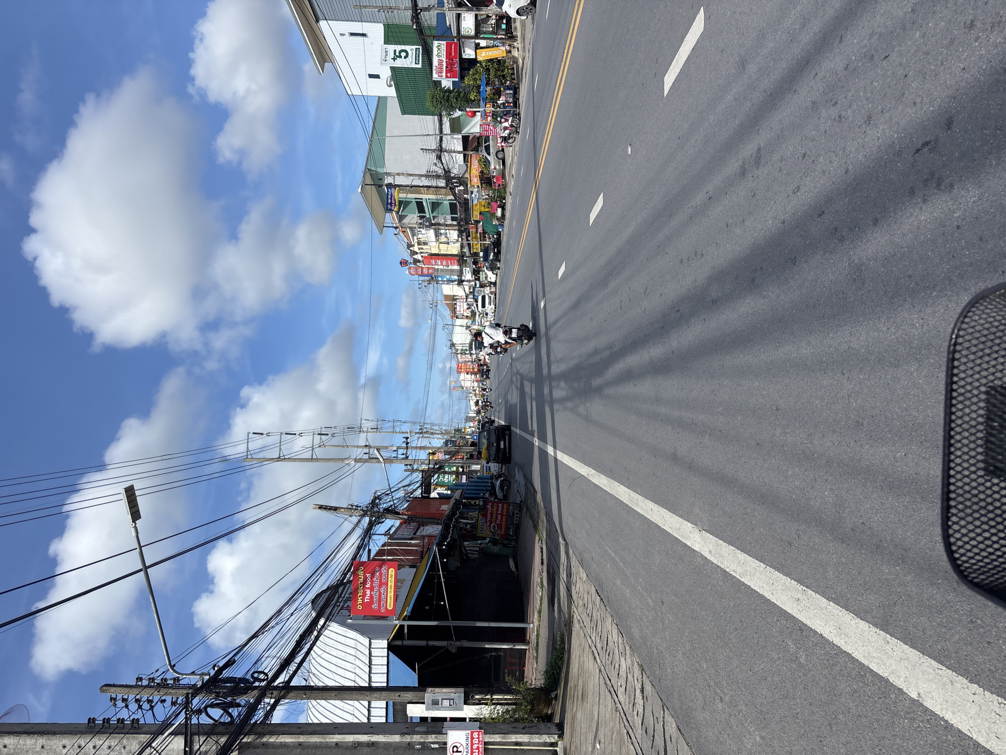 The image depicts a street scene in Chalong, Thailand, under a bright blue sky with scattered white clouds. The perspective is from a moving vehicle, possibly a scooter, looking forward along a two-lane asphalt road. On the left side of the road, there are buildings with awnings and a sign in Thai script advertising "Thai food" and "Noodles soup." A "P" parking sign is also visible on the left. The right side of the road features a row of multi-story buildings, some with balconies and air conditioning units. One building has a green facade with the number "5" and the word "Pharmacy" on it. Numerous utility poles with a dense network of electrical wires are prominent along both sides of the street. Several motorcycles and a few cars are present on the road, with some people riding on motorcycles. The sun is casting long shadows across the asphalt. The overall impression is a typical bustling street in a Thai town.