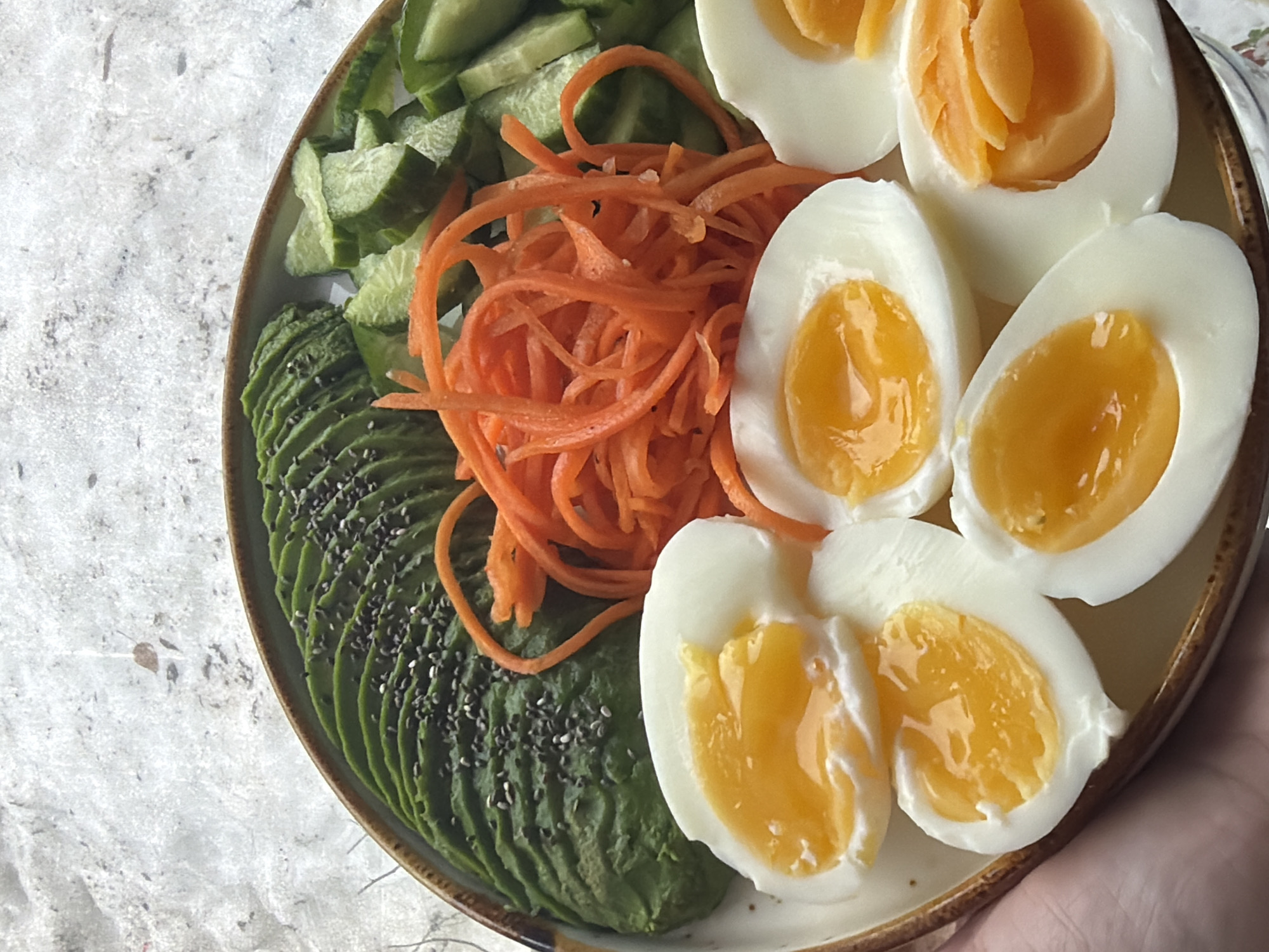 A high-angle, close-up photograph displays a meal served on a round, light-colored ceramic plate with a textured brown rim. A human hand is partially visible in the lower-left quadrant, holding the plate. The plate's contents include several food items: fanned slices of green avocado, sparsely topped with black chia seeds, positioned in the upper-left. Adjacent to the avocado are bright orange, julienned carrots. In the upper-right, chunky, roughly chopped green cucumber pieces are arranged. Across the bottom and center, multiple halves of soft-boiled eggs are visible, showcasing solid white albumen surrounding vibrant orange-yellow, semi-liquid yolks. The background is a light gray, mottled surface, consistent with a stone countertop or table. The specific location within Perm, Russia, cannot be determined from the visual information present in the image.