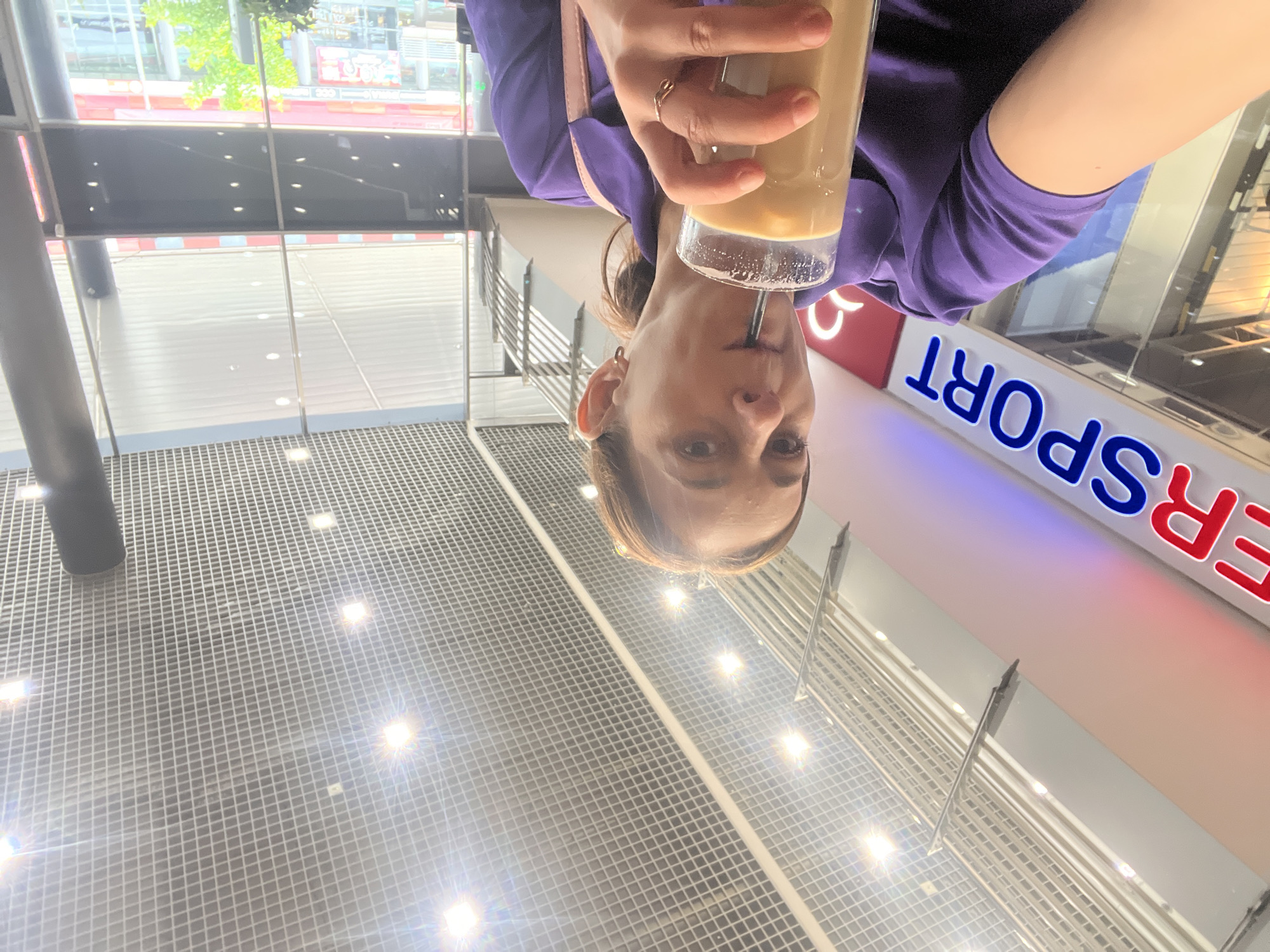 A woman in a purple shirt is holding a glass of iced coffee and drinking it through a straw. She is wearing earrings and a ring. Behind her is a sign that reads "ER SPORT". The ceiling is made of a grid of lights and metal. The environment appears to be an indoor shopping center or mall.