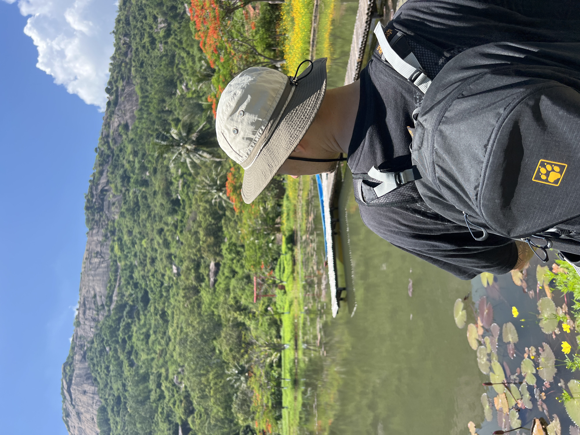 A person wearing a black t-shirt, a light-colored bucket hat, and a black backpack is standing and looking out at a lake. The lake has lily pads and yellow flowers along its edge. In the background, there is a dense green landscape with a large rocky mountain and trees with orange flowers. A wooden dock extends into the lake. A red torii gate is visible in the distance among the trees. The sky is blue with a large white cloud. The location is identified as Nam Cam Ranh, Vietnam.