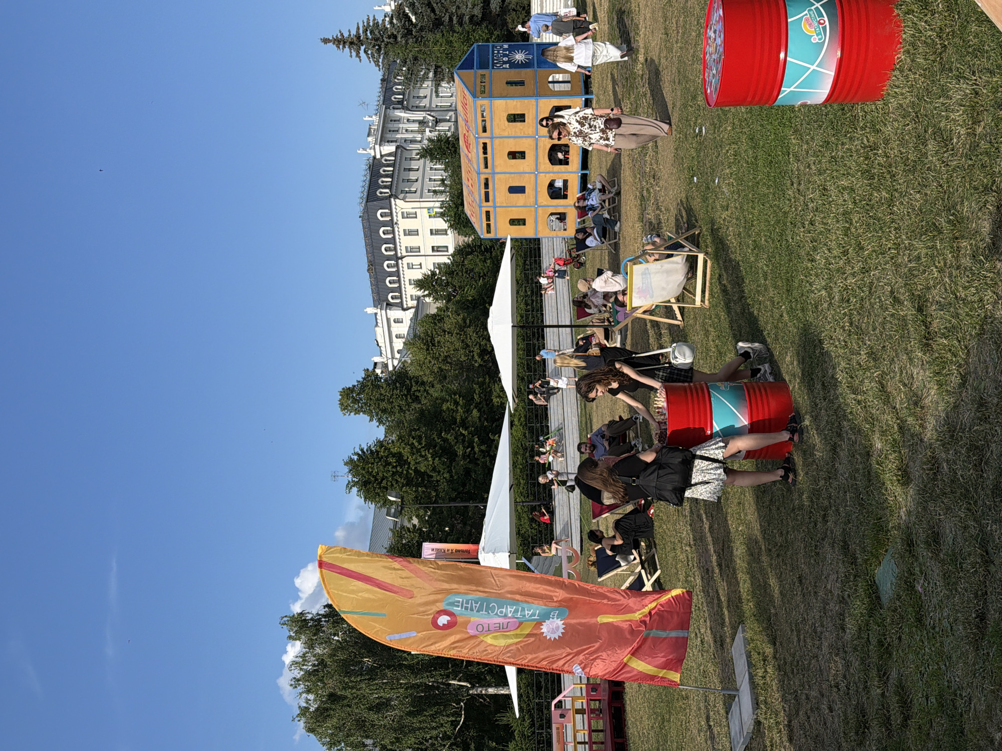 The image depicts an outdoor event taking place in Kazan, Russia, likely on a sunny day with clear blue skies. The scene is set on a grassy lawn with several people enjoying the festivities. In the foreground, two women are standing next to a stack of red barrels, one of which has a graphic design and text that appears to be in Russian. A large, colorful orange flag with graphic elements and text is prominently displayed to the left of the frame.
Scattered across the lawn are lounge chairs, with several individuals relaxing in them. In the middle ground, a distinctive structure resembling a multi-story building with an orange exterior and blue accents stands out, possibly serving as an event pavilion or display. White umbrellas provide shade for some areas of the event.
In the background, a grand building with a light-colored facade and dark rooflines, likely a historical or public building in Kazan, overlooks the event. Trees and greenery surround the area, contributing to a park-like atmosphere. People are seen walking, sitting, and interacting, suggesting a casual and social gathering. The overall impression is of a vibrant public event in an urban setting.