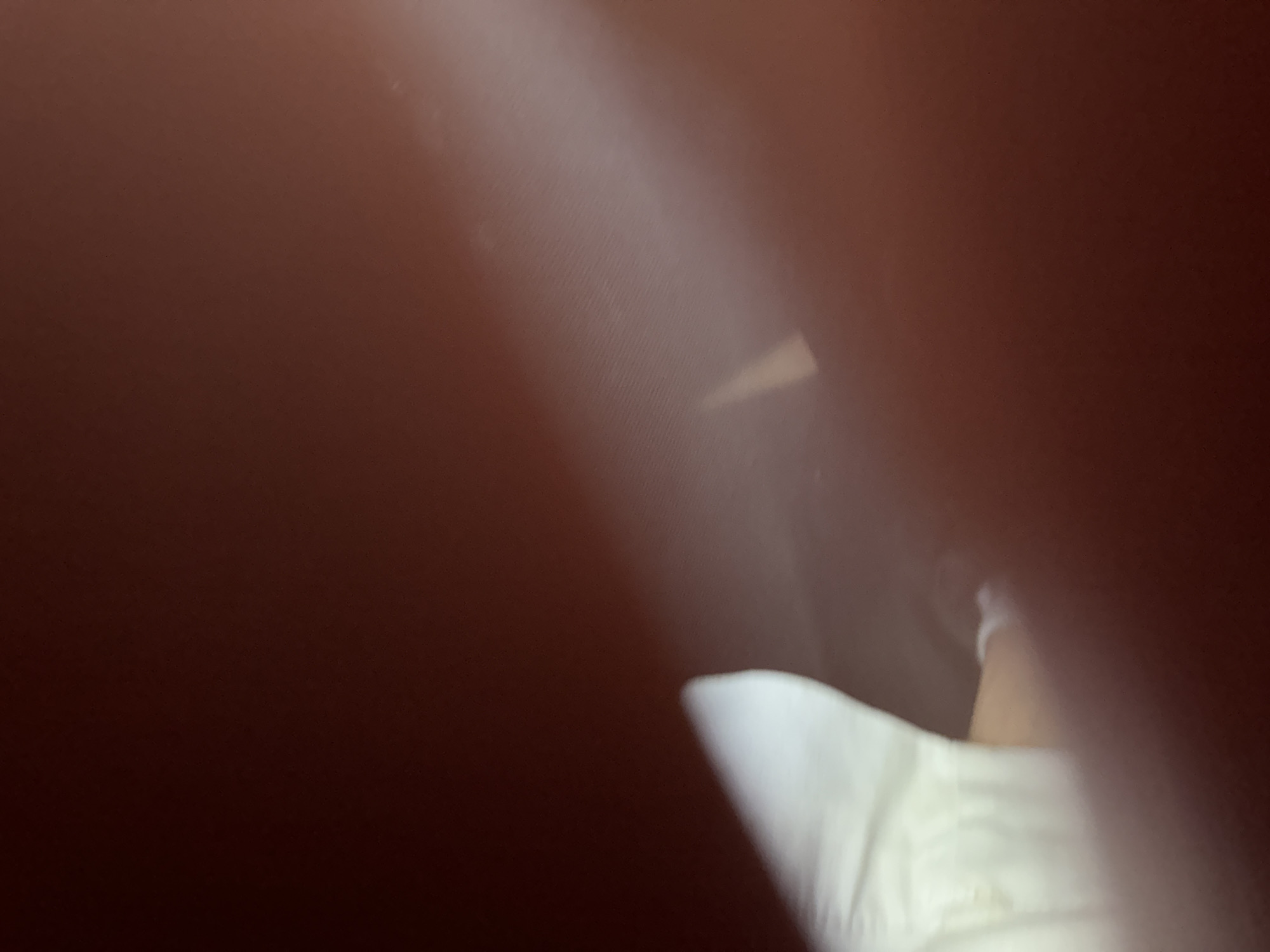 The image shows a blurry close-up of a white object, possibly fabric or a sleeve, in the lower left quadrant, illuminated by a bright light source originating from the upper right. The background is a dark reddish-brown, with a diffused beam of light cutting across the scene diagonally. No specific location in Varna, Bulgaria can be identified from the visual information. The image lacks any identifiable people, objects, or actions, and the environment is indeterminate.