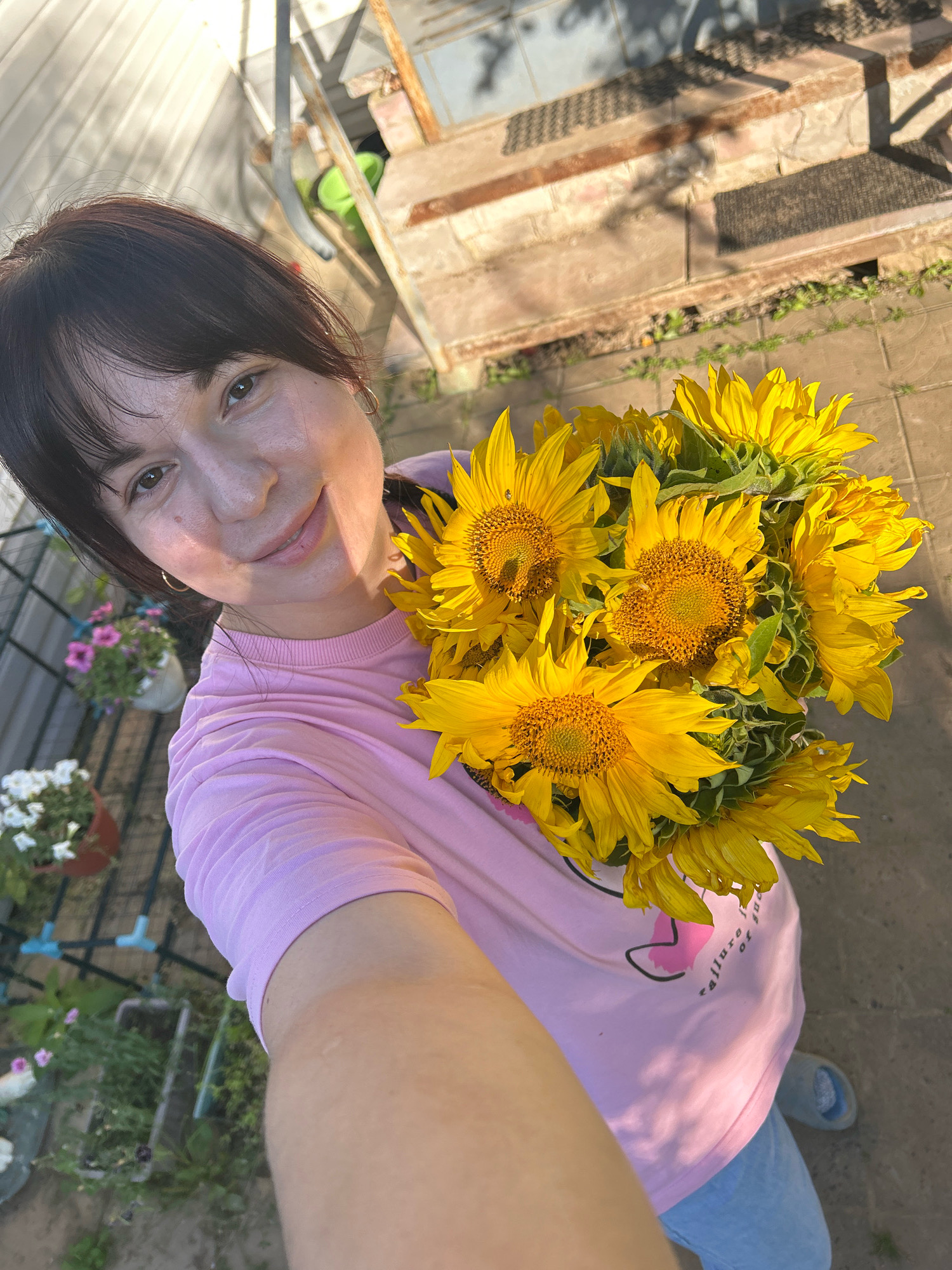 A woman in a pink t-shirt and light blue pants is holding a bouquet of sunflowers. She is outdoors, with a paved ground visible and some greenery and potted plants in the background. Behind her, there is a structure that appears to be a greenhouse or shed with metal and wooden elements. The scene is illuminated by sunlight, casting shadows on the ground. There are no visible landmarks in the image that would specifically indicate the location in the city of Фоки, Russia.