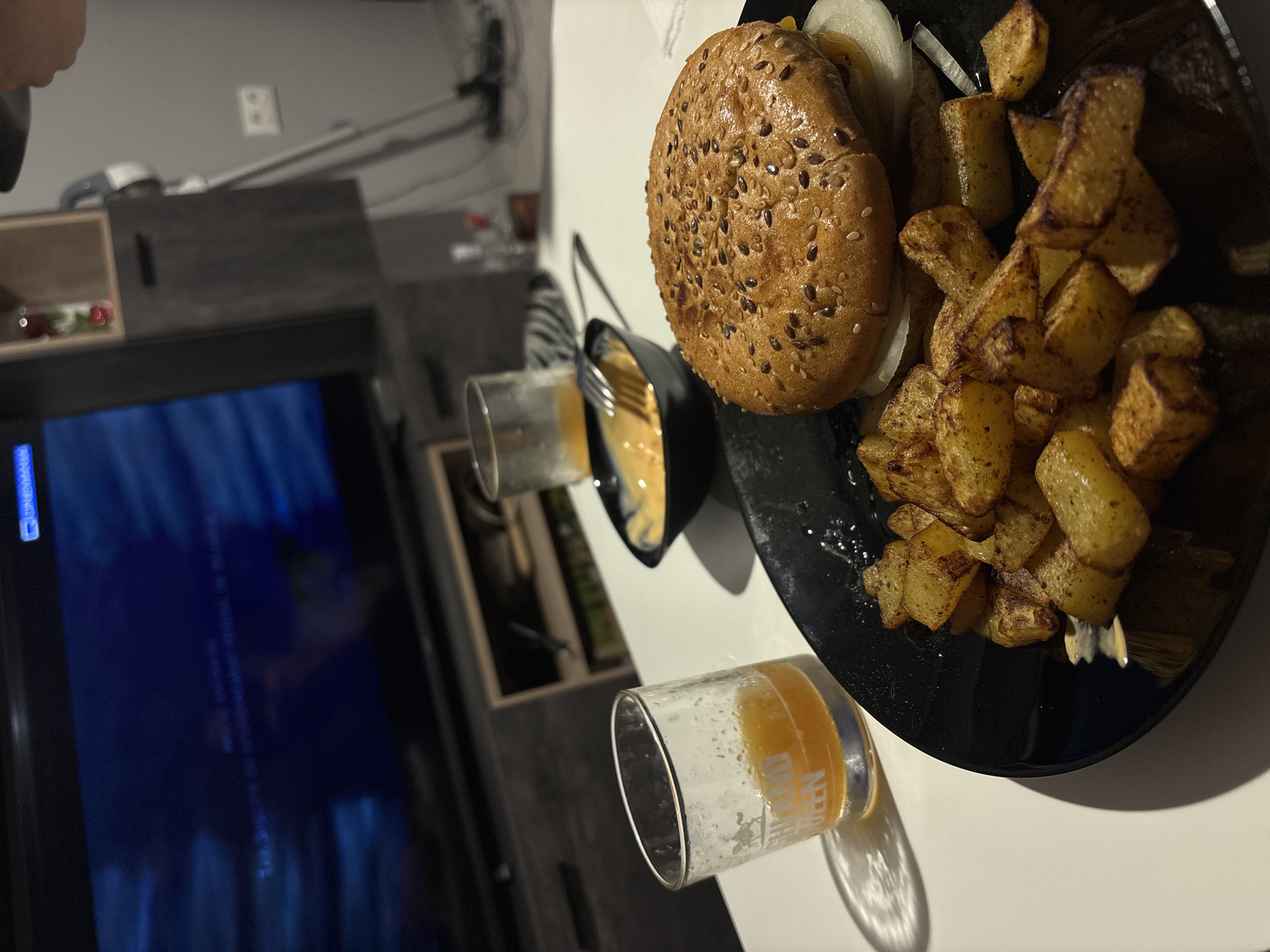 The image presents an indoor dining scene, likely during the evening or night, set in what appears to be a living room or dining area in Varna, Bulgaria. On a white surface, a black plate holds a meal consisting of a burger with a sesame and flaxseed seed bun, revealing slices of white onion. Beside the burger is a generous serving of seasoned potato wedges or fries. A small black bowl containing a light yellow sauce, possibly cheese or mayonnaise, with a fork inside, is also part of the meal.
Two clear glasses are visible. One glass, containing a light orange beverage (possibly beer or juice), has the text "BRAND QUEEN" and other indistinct writing printed on its side. The second glass is mostly empty, showing residual liquid.
In the background, a large television screen displays abstract blue and dark patterns. Faintly visible in the upper left corner of the screen is the word "WARNING." Below the TV are dark-colored modular shelves or cabinets. A light-colored wall with a white electrical outlet is seen higher up. A person's hand is partially visible in the top left of the frame. The lighting is dim and artificial, casting shadows, and the overall scene suggests a casual meal at home while relaxing and watching television.