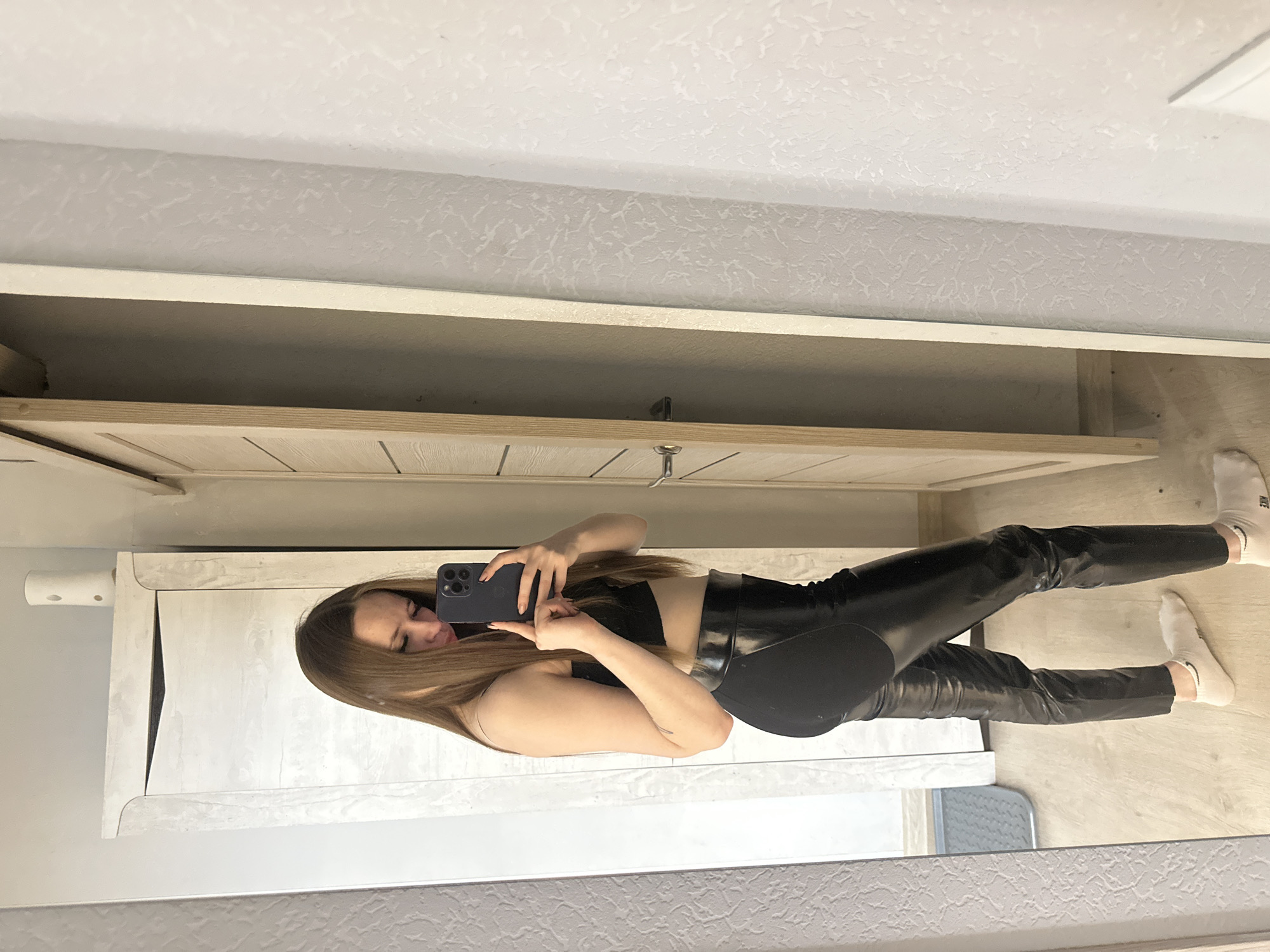 A woman is taking a selfie in a bathroom. She is wearing a black crop top and black leggings. She is standing in front of a mirror that is attached to a vanity with drawers. The bathroom floor is light colored wood and the walls are light colored.