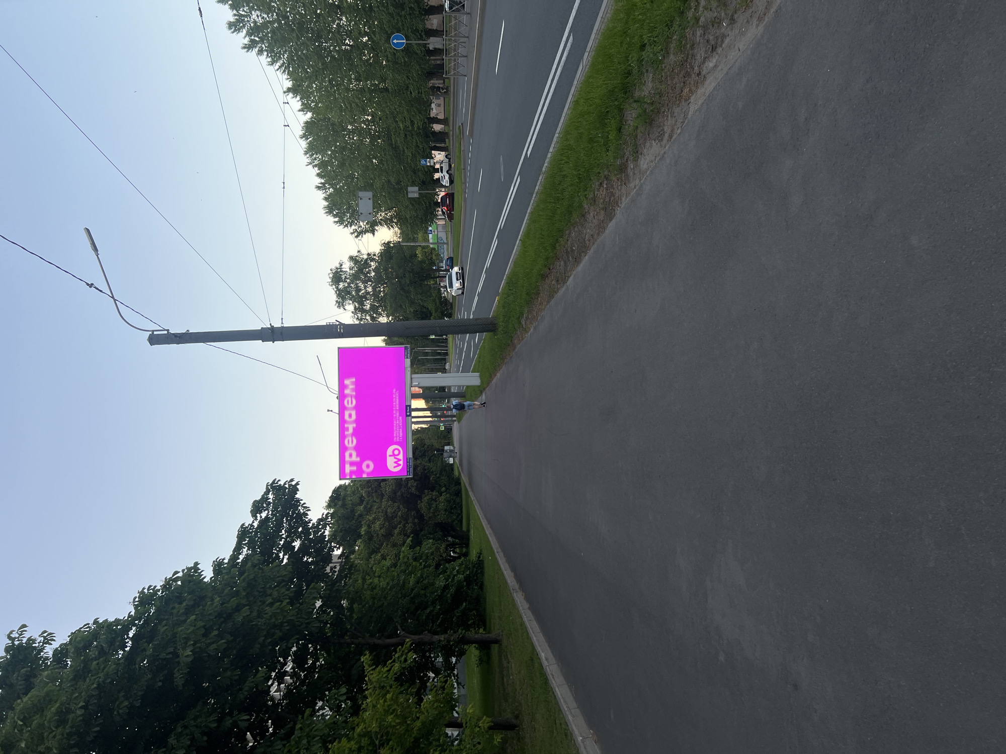 The image displays an urban street scene under a clear, light blue sky, likely in the late afternoon or early evening in the city of Unknown, Russia. A wide asphalt sidewalk runs along the left side of the frame, bordered by a concrete curb and a dense line of mature green trees. On the right, a multi-lane asphalt road with white lane markings stretches into the distance, carrying several vehicles.
A prominent feature is a large, brightly illuminated digital billboard positioned on the right side of the sidewalk. The billboard displays a vibrant magenta background with white Cyrillic text, reading "стречаем" (strechaem, meaning "we meet" or "we welcome") on the first line, followed by a partially visible "о" on the second. Below the text, a white "wb" logo, indicative of Wildberries, is enclosed within a purple circle, accompanied by smaller, unreadable text. The billboard is supported by a dark grey pole, which also anchors a utility pole extending upwards with a streetlight arm and overhead wires.
On the far right of the sidewalk, a single individual is walking away from the camera, wearing a light-colored top, dark shorts, and a backpack. On the road, a white sedan is visible in the left lane, driving towards the background. Further down the road, a dark red SUV and a white SUV are either parked or slowly moving. Traffic signs, including a blue circular sign with a white arrow (indicating 'forward only') and a 'P' for parking, are visible on the far side of the road. More trees line the opposite side of the road, providing a green backdrop to the scene. The ground surface of both the sidewalk and the road appears to be dark, worn asphalt.