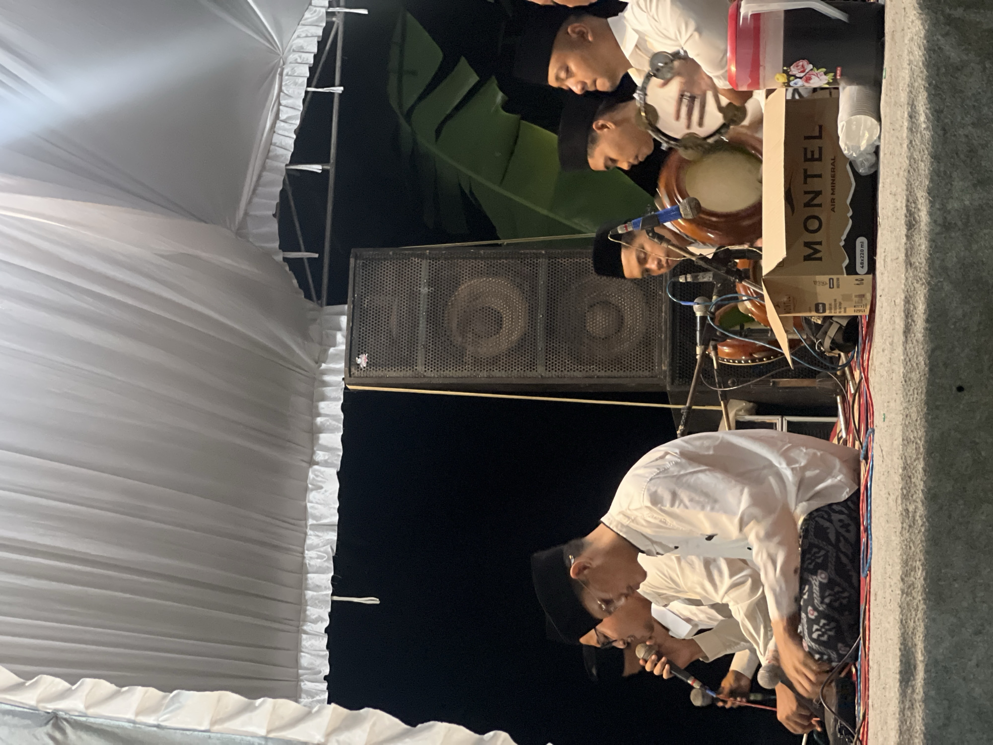 This image captures a group of young men performing traditional Indonesian music under a white tent. The setting appears to be outdoors, possibly during an evening event, given the dark background and artificial lighting. Several individuals are seen playing musical instruments, including drums and what appears to be a microphone, suggesting a musical performance or religious gathering. They are all dressed in traditional attire, featuring white shirts and black skullcaps, a common practice in Indonesia for religious or cultural events. In the foreground, a cardboard box labeled "MONTEL AIR MINERAL" is visible, along with disposable cups, indicating refreshments are available for attendees. The overall atmosphere seems to be one of communal gathering and cultural celebration. This event is taking place in Wedarijaksa, Indonesia, a region known for its rich cultural heritage and traditional music.