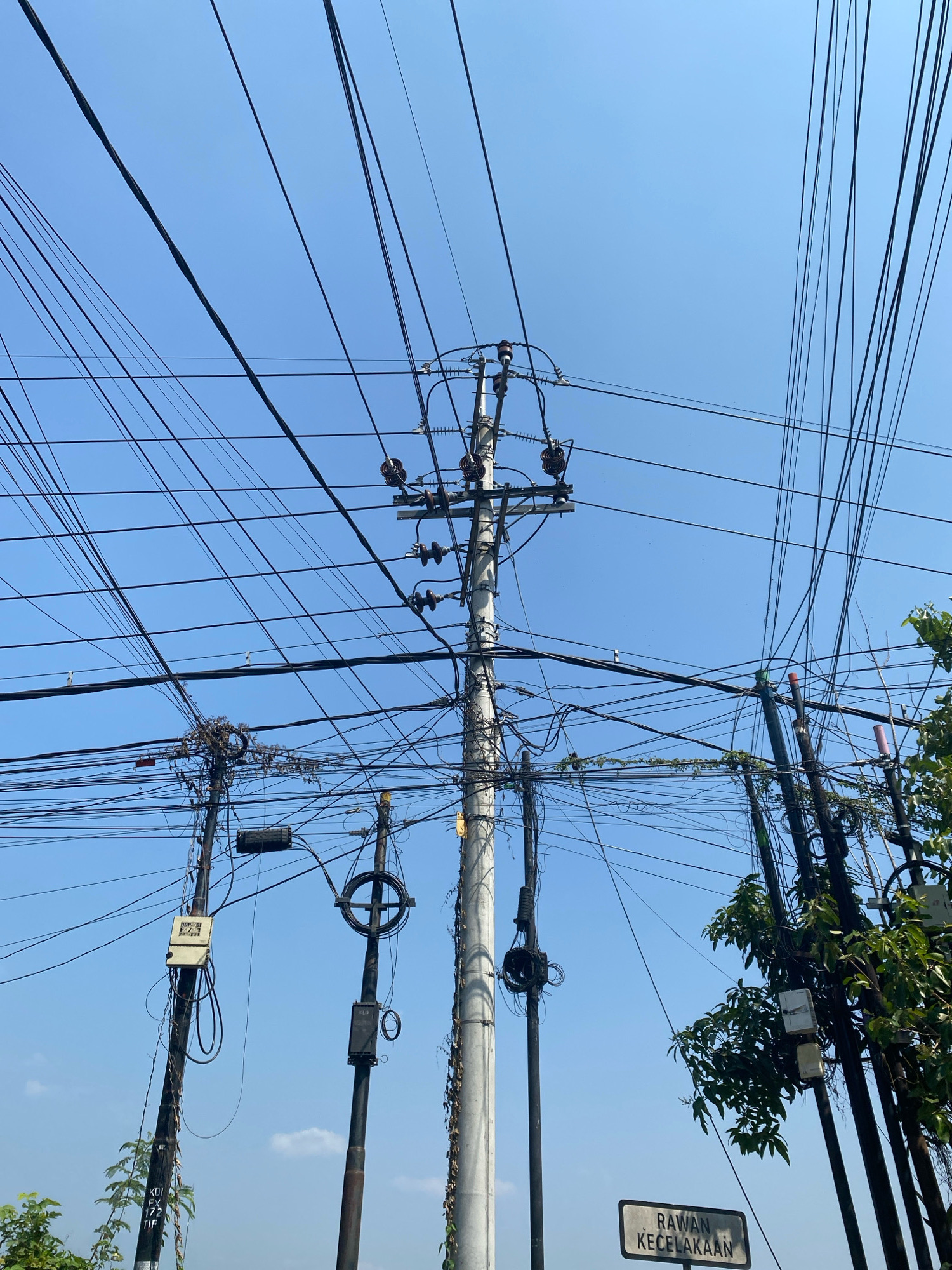 A low-angle, wide shot captures a dense network of overhead power lines and telephone wires against a clear blue sky. Multiple utility poles, constructed from concrete and metal, are visible. Some poles are adorned with electrical insulators and transformers. Various electrical boxes and junction boxes are attached to the poles, with cables running in all directions, creating a chaotic and tangled appearance. Vegetation, including vines, grows on some of the poles and wires. A sign with the text "RAWAN KECELAKAAN" is mounted on a pole in the lower right portion of the frame. The image suggests an urban or suburban environment in Kediri City, Indonesia, likely a street or intersection characterized by extensive utility infrastructure. There are no people or animals visible in the scene.