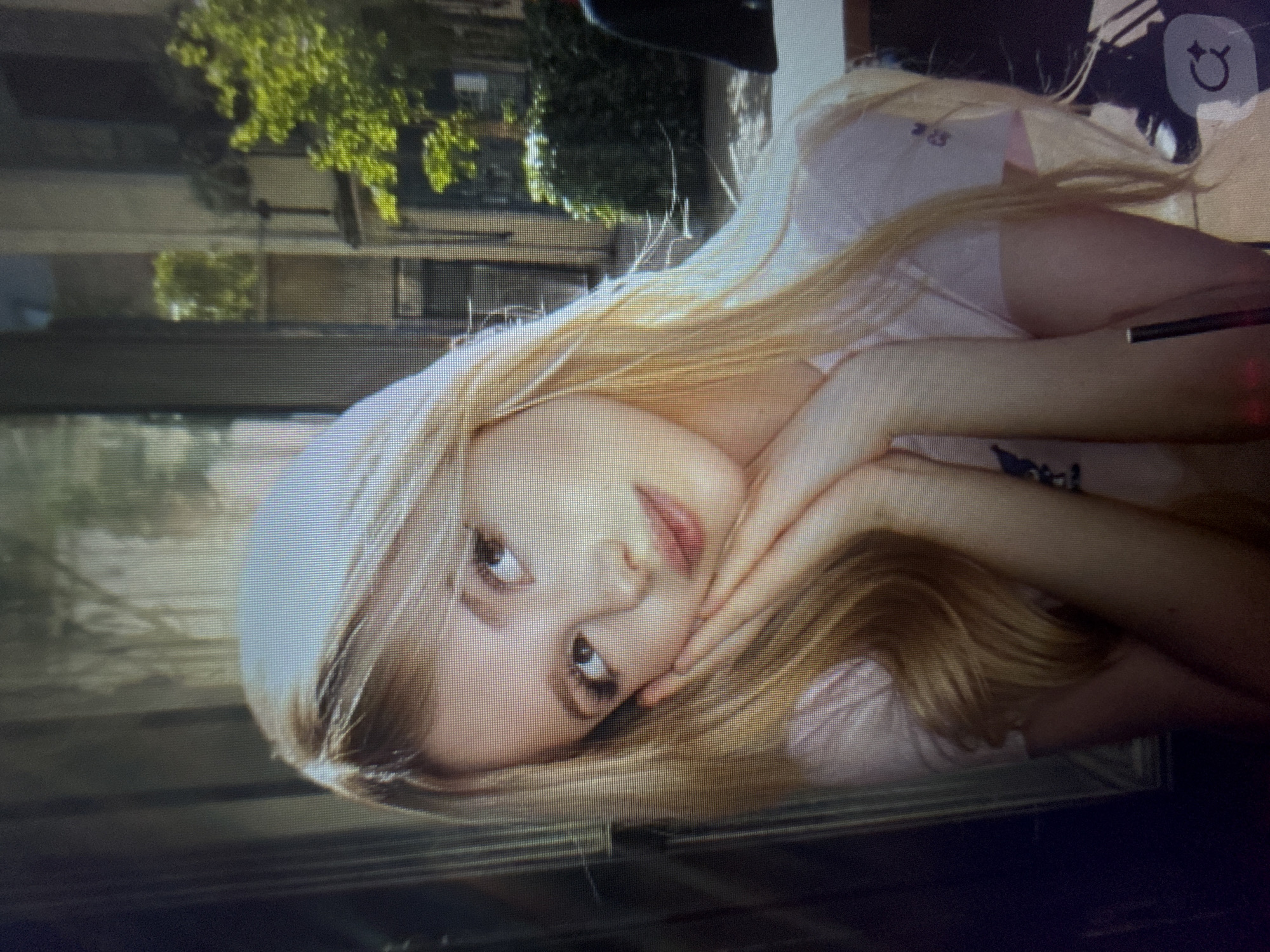 A full-body photograph, taken of a screen, depicts a young woman with long, straight blonde hair and light skin, rotated 90 degrees counter-clockwise. Her head is positioned towards the center-left, with her gaze directed towards the viewer. She is resting her chin and cheek on her interlocked hands, creating a contemplative or posed expression. Her lips are slightly parted, revealing a hint of a pink tint.
She wears a light-colored top, possibly white or pale pink. A faint, small number "25" is visible on the upper part of her left arm/shoulder. Near her right arm, a dark, thin object resembling a pen or stylus is present. The entire image exhibits a visible pixel grid pattern, characteristic of a photograph taken of a digital display.
The background, partially out of focus, shows an outdoor setting with green foliage, suggestive of trees or bushes, and muted architectural structures, indicating an urban or suburban environment. The lighting appears to be natural daylight, casting highlights on her hair and face. In the top right corner of the image, a white digital icon, possibly from a social media or camera application, depicts a smiling face with sparkling eyes and a downward-pointing arrow. The overall impression is a casually posed portrait captured from a screen.