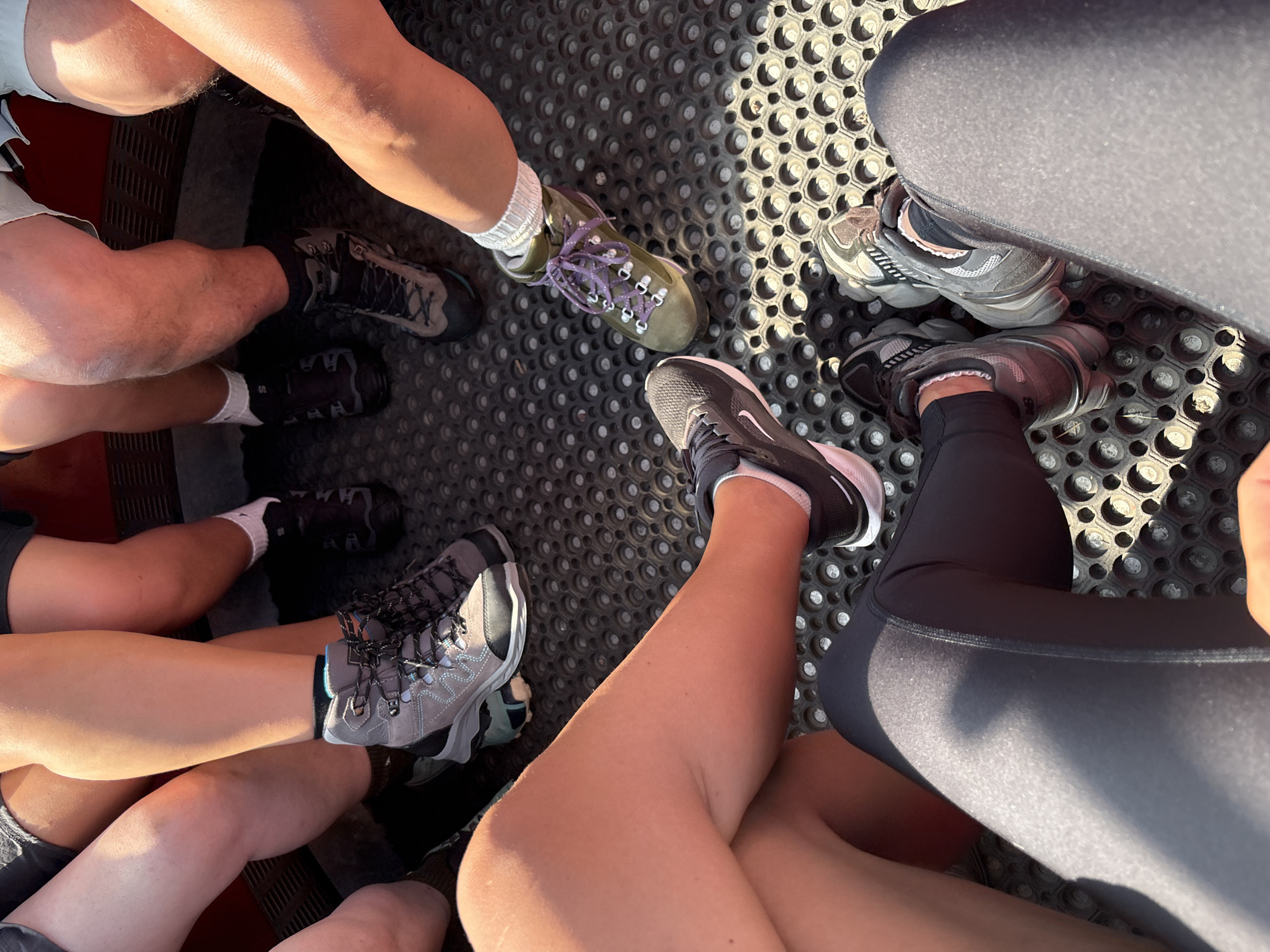 In this overhead, close-up shot, four pairs of legs and feet, clad in various hiking boots and athletic shoes, are positioned around a circular, dark-colored mat with a honeycomb pattern. The lighting suggests it is daytime, casting shadows and highlighting the sun-kissed skin of the individuals. The setting appears to be outdoors, possibly in a natural or recreational area within the city of Kastelruth - Castelrotto, Italy, given the presence of hiking attire. The individuals are not fully visible, with the focus being on their lower extremities and footwear, implying a casual gathering or a break during an outdoor activity. The perspective suggests the photographer is also part of this group.