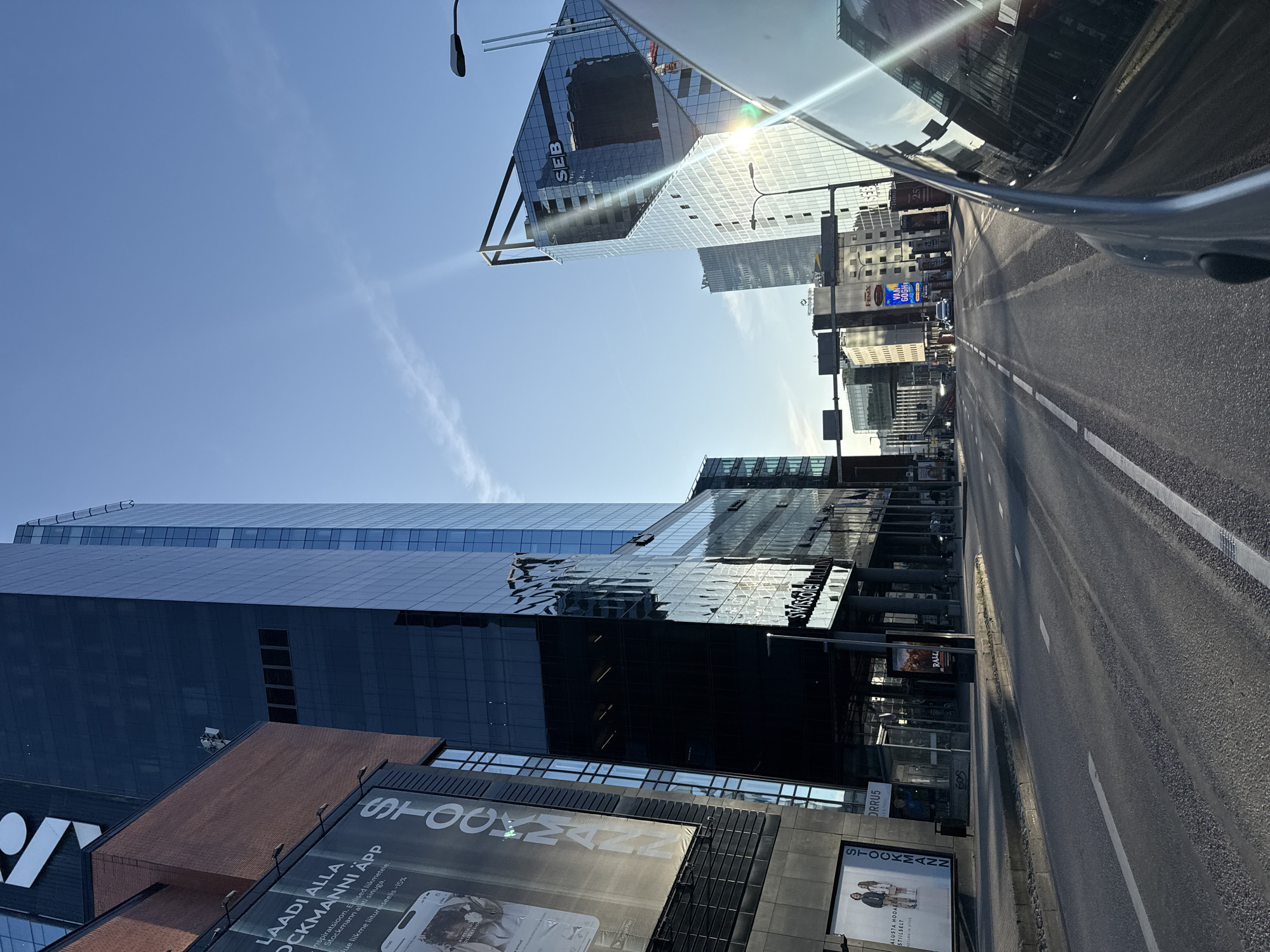 The image captures a street view in Tallinn, Estonia, looking towards modern glass-fronted buildings. The prominent structures include the Stockmann department store building on the left and the SEB bank building on the right, identifiable by its logo. The scene is dominated by the clear blue sky and the sun, which creates lens flare across the right side of the image, reflecting off the car's windshield from which the photo is taken. The street itself is paved and marked with white lane lines. Several vehicles are visible on the road, including cars and what appears to be a bus or tram. Advertisements are displayed on billboards and building facades, including one for "STOCKMANN APP" and another featuring a "VAN GOGH" exhibition. The overall impression is of a contemporary urban environment bathed in bright daylight.