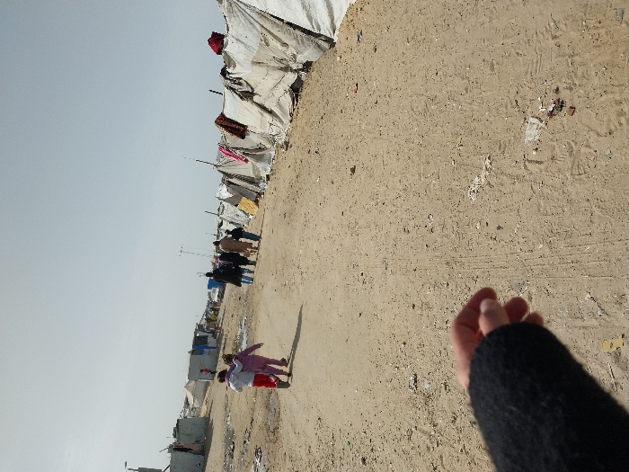The image depicts a makeshift settlement in Khan Yunis, Palestinian Territory, under a hazy sky. An unpaved, sandy dirt path forms the central axis, showing faint tire tracks and scattered litter. In the foreground, the lower left displays a person's left hand and forearm, clad in a black, textured sleeve, extending into the frame from the perspective of the photographer.
In the midground, two young children walk away from the camera; one wears a red top with light pants, and the other wears a light purple full-body suit. Further along the path, a group of approximately five adults and one child also walk away, dressed in various garments.
To the left of the path, several rudimentary shelters are constructed from corrugated metal and other salvaged materials, surrounded by debris. To the right, a continuous line of temporary shelters, primarily made from white and light-colored tarpaulins or fabrics draped over frames, extends into the background. Several colorful blankets and garments are draped over these tents. The distant background shows additional makeshift structures and utility poles.