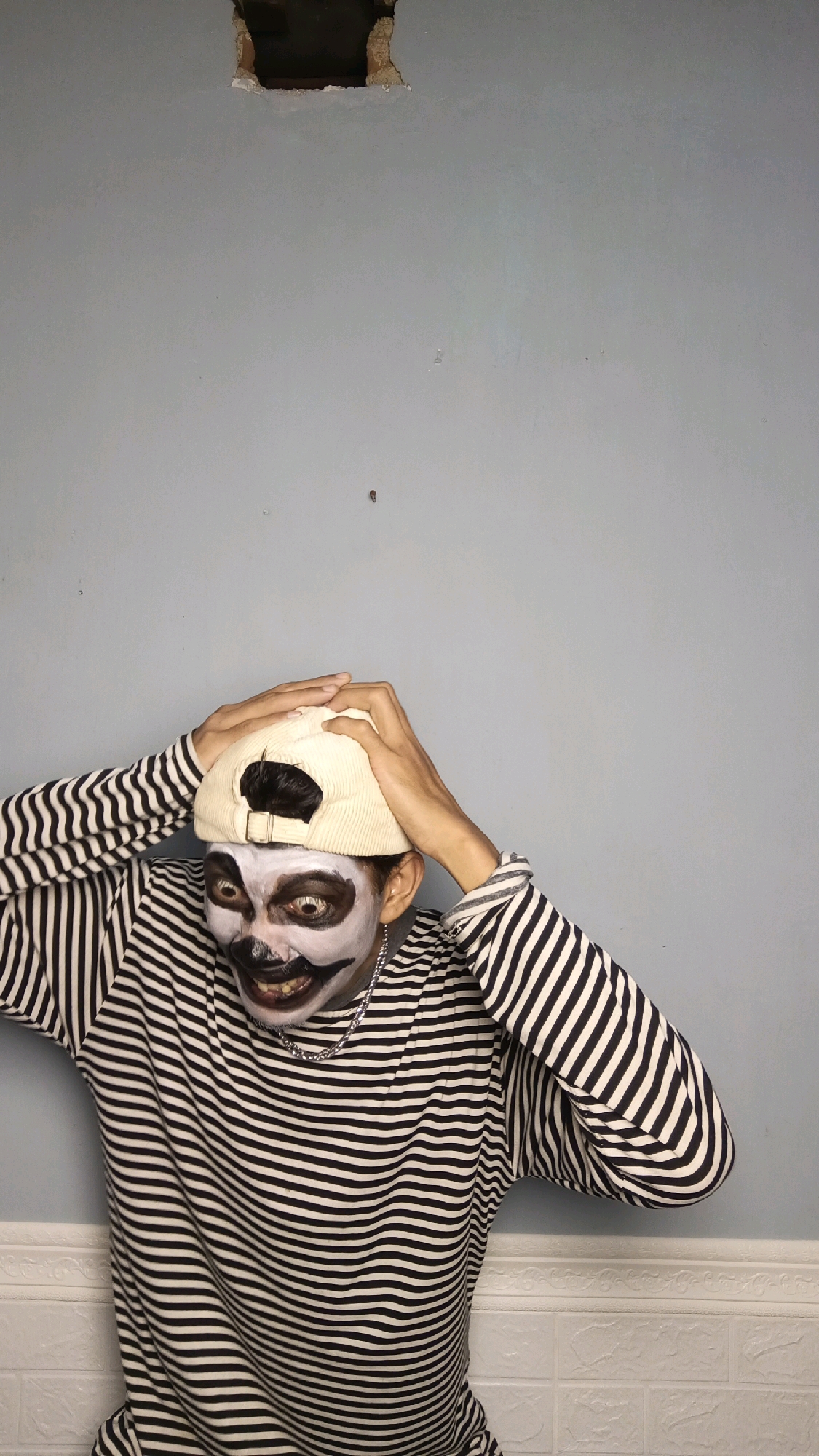 In a studio setting, a person with clown makeup, featuring exaggerated black and white features and dark lipstick, is captured from the chest up. They are wearing a long-sleeved black and white striped shirt and a light-colored baseball cap, which they are adjusting with both hands. The background consists of a light blue wall with a rectangular hole near the top, revealing darker material behind it, and a white brick-patterned wainscoting along the bottom. The lighting is even, highlighting the subject's makeup and attire. No specific location in Kebumen, Indonesia is discernible from the image.