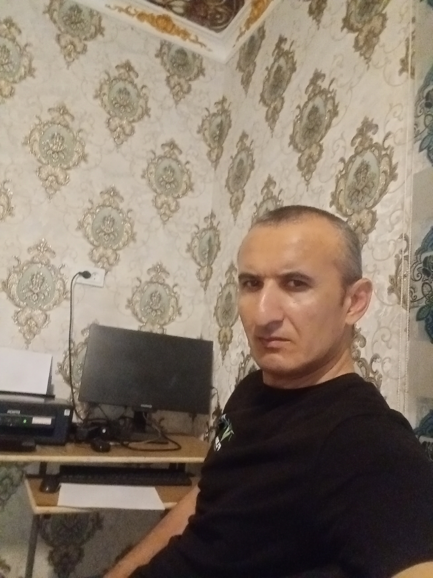 A man with short, dark hair and a serious expression is seated in front of a desk in a room. He is wearing a black t-shirt. On the desk are a computer monitor, a printer, and a keyboard. The walls are covered with a patterned wallpaper featuring ornate medallions. There is also an electrical outlet visible on the wall. The image appears to be a selfie taken indoors. The location is identified as the city of Karagana, Uzbekistan.