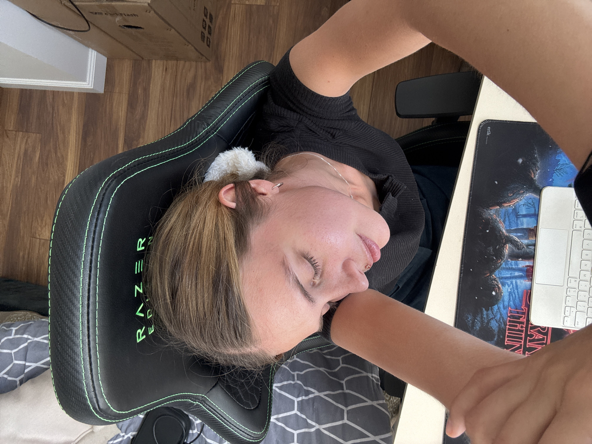 An overhead shot captures a person, appearing to be a young woman, resting her head on her right arm with her eyes closed, suggesting sleep or deep relaxation. She is seated in a black Razer gaming chair, identifiable by the "RAZER" logo and neon green stitching on the headrest. Her light brown hair is partially pulled back with a white fluffy scrunchie, and she wears a black ribbed long-sleeved top, a silver chain necklace, and has an ear piercing and a small stud on her upper lip.
The chair is positioned at a light-colored desk, where a large mousepad is visible. The mousepad features a "Stranger Things"-themed design with dark, monstrous figures against a blue, wintery background. The text "STRANGER THING" is partly visible in red on the mousepad, along with a small tag reading "ERIK". A partial view of a white keyboard, likely an Apple Magic Keyboard, is also on the desk.
The background shows a warm-toned wooden plank floor, a white baseboard, and a partially visible cardboard box. To the lower left, a grey and white geometric patterned fabric, possibly a blanket or duvet cover, is seen. The scene depicts a quiet indoor setting, likely a personal room or office.