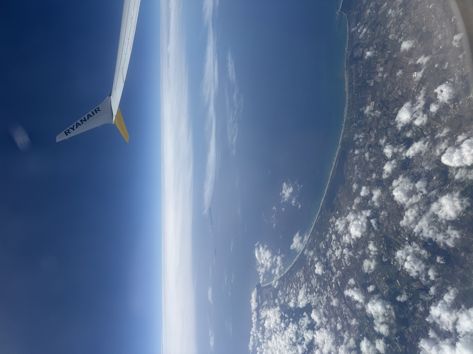 The image is taken from inside an airplane, likely a Ryanair flight based on the visible wing. The wing is white with the "RYANAIR" logo in blue and a gold-colored tip. Looking out the window, there's a clear view of the Earth. A coastline is visible, with a long, sandy beach bordering the ocean. Inland from the beach, the landscape is a mix of fields and urban areas, partially obscured by scattered white clouds. The ocean is deep blue, transitioning into lighter shades as it approaches the horizon, which is somewhat hazy. The sky above is also blue, fading to lighter shades toward the horizon.