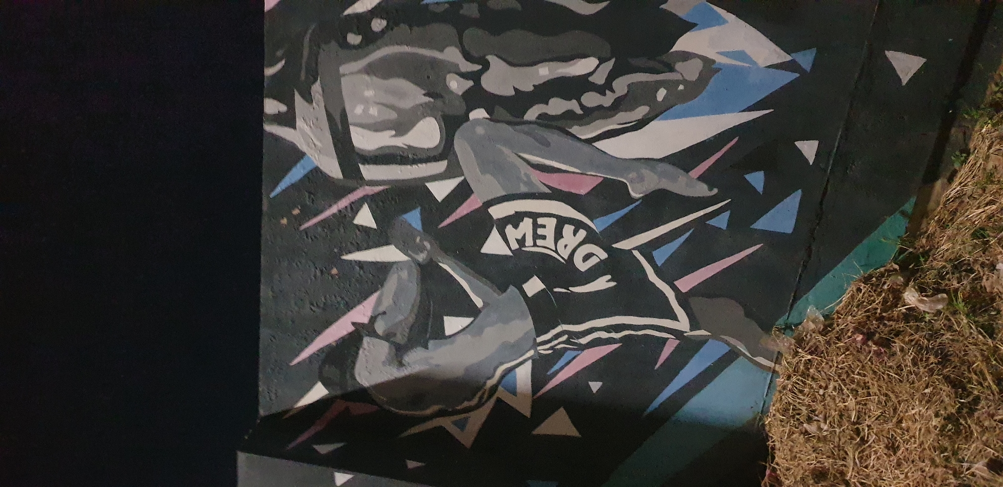 A mural depicts a stylized representation of a Muay Thai fighter in action, rendered in grayscale with accents of pink, blue, and white. The fighter, depicted from the torso down, is captured mid-kick, with one leg extended forward and the other bent at the knee. They are wearing shorts with white stripes and the word "DREW" emblazoned across the thigh. Behind the fighter, abstract geometric shapes in pink, blue, and white create a dynamic, energetic background against a dark, textured surface, likely a wall. To the right, partially obscured, is a shadowy figure, possibly an opponent or a thematic element, also rendered in grayscale with swirling, abstract details. The lower portion of the mural is obscured by a pile of dry, straw-like material and some scattered pieces of clear plastic, suggesting an outdoor urban environment. This scene is located on a wall in the city of Narayi, Nigeria.