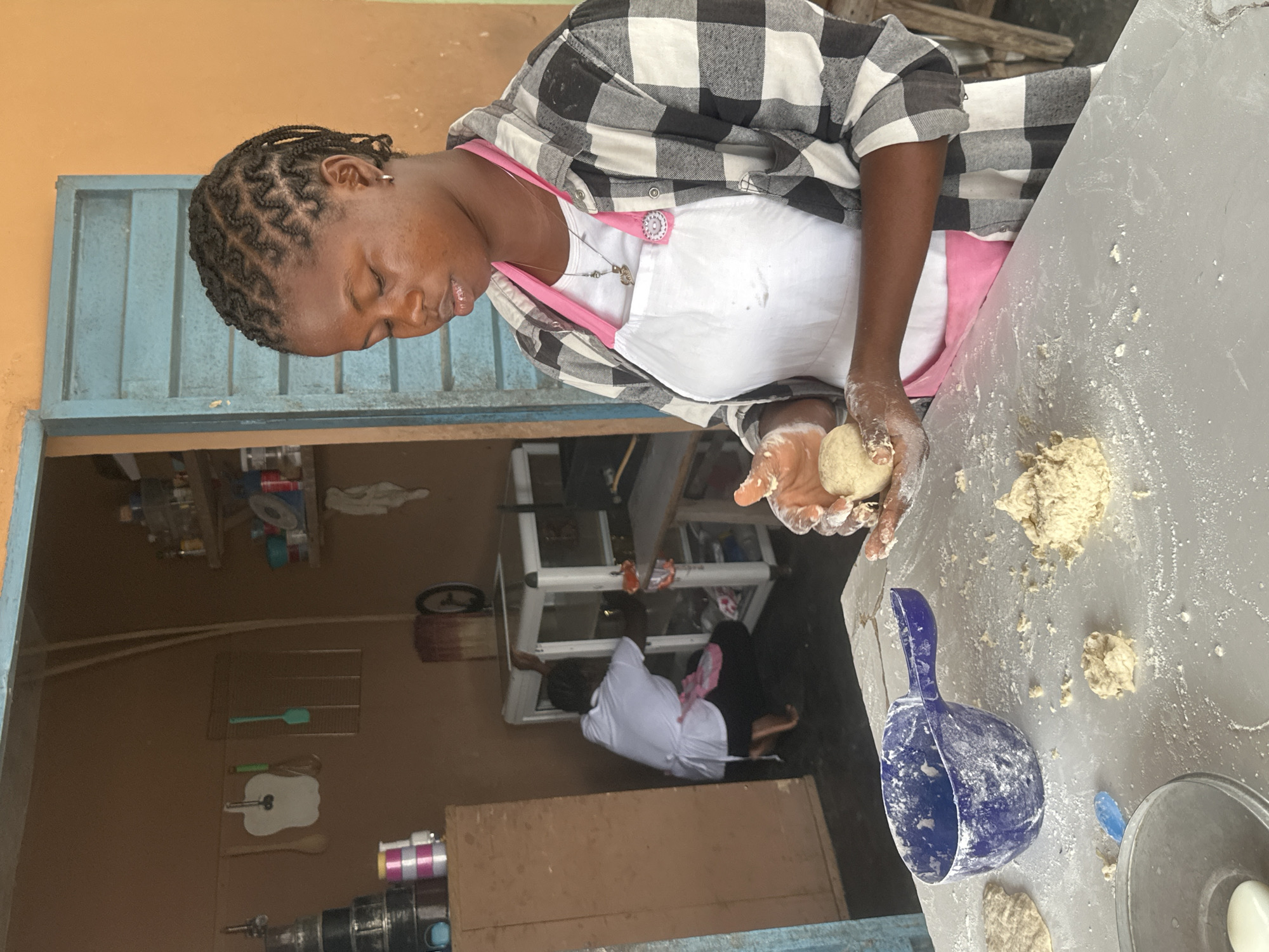 A young woman, with her dark hair in braids, is kneading dough on a flour-dusted table in what appears to be a kitchen or food preparation area. She is wearing a pink apron over a white t-shirt and a black and white checkered shirt. She holds a ball of dough in her hands. A blue plastic scoop, a metal bowl, and scattered pieces of dough are visible on the table. In the background, a person in a white shirt and apron is crouched near a glass-fronted cabinet. Shelves with various items, kitchen utensils, and cooking equipment are visible in the room. The environment suggests a modest, functional space, possibly a small business or community kitchen in Badagry, Nigeria.