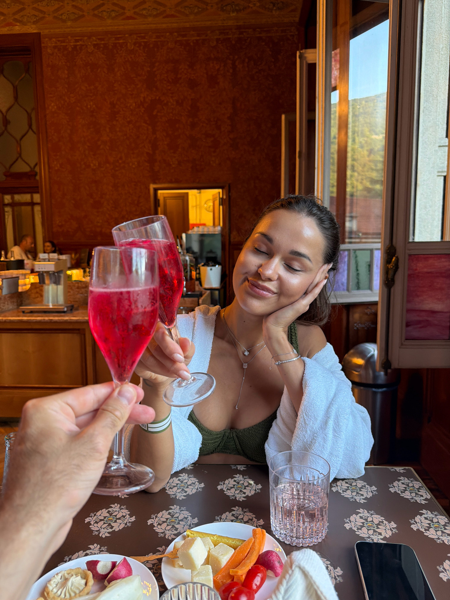 The image depicts a woman with her eyes closed, resting her cheek on her hand, with a content expression. She is wearing a green bikini top and a white towel draped over her shoulders. She is holding a champagne flute filled with a pink beverage, toasting with another person whose hand and arm are visible in the foreground, also holding a similar flute. In front of them, on a table with a patterned tablecloth, is a plate of cheese and vegetables, a glass of water, and a smartphone. The background suggests an indoor setting with ornate wallpaper and a window revealing a view of trees and a building. The woman's jewelry, including necklaces and bracelets, is visible. The overall scene conveys a relaxed and celebratory atmosphere, possibly at a spa or a resort in San Pellegrino Terme, Italy, given the context provided.