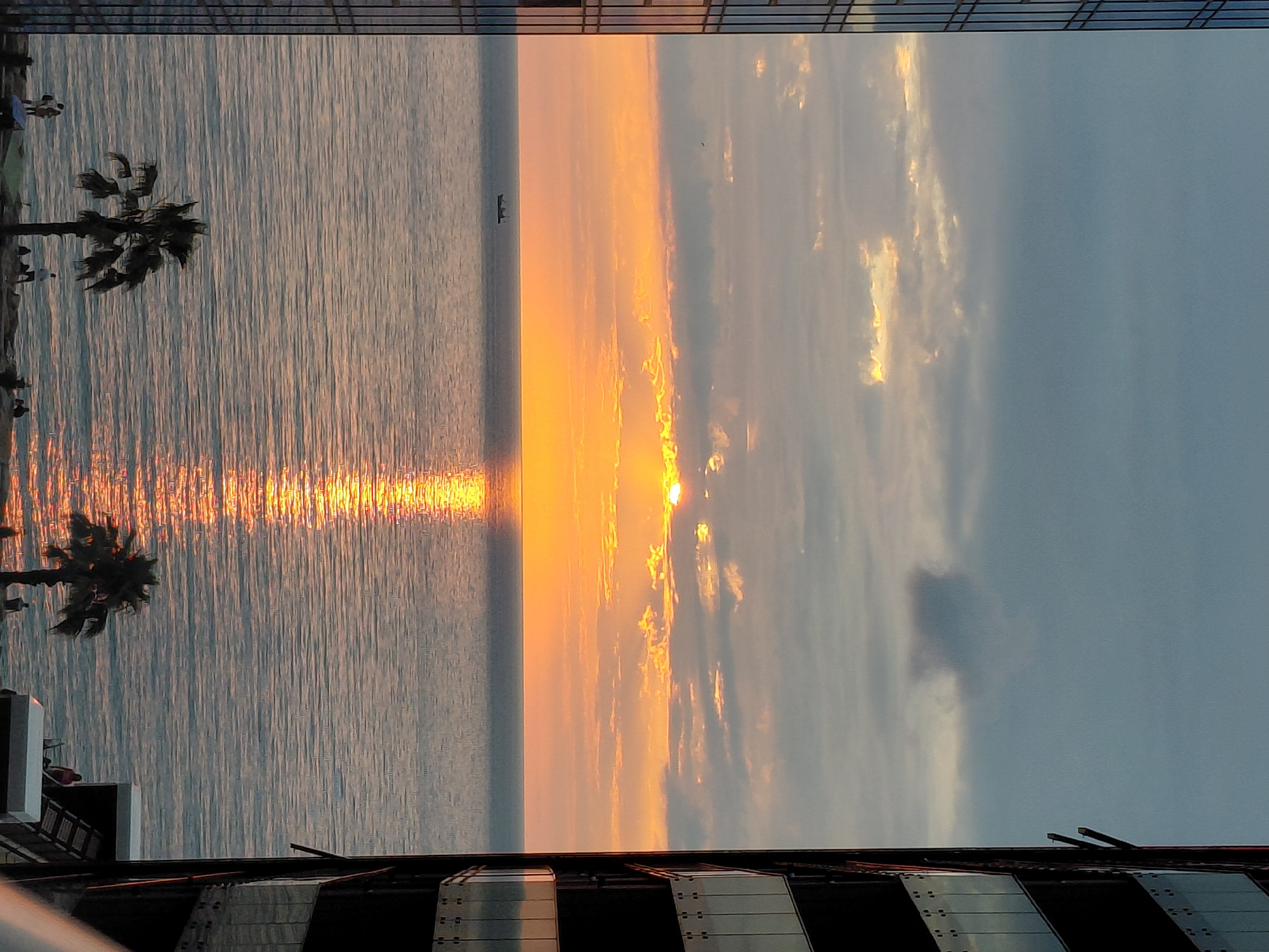 A sunset over the ocean is depicted, framed by the sides of two modern buildings. The sun is low on the horizon, casting a bright orange glow that reflects on the water. Clouds are present in the sky, some tinged with the colors of the sunset. A small boat is visible on the water. In the foreground, palm trees and a few silhouetted figures can be seen on the shoreline, suggesting a beach or promenade area. The buildings have glass facades, and balconies are visible. The overall scene is one of a coastal city at dusk.