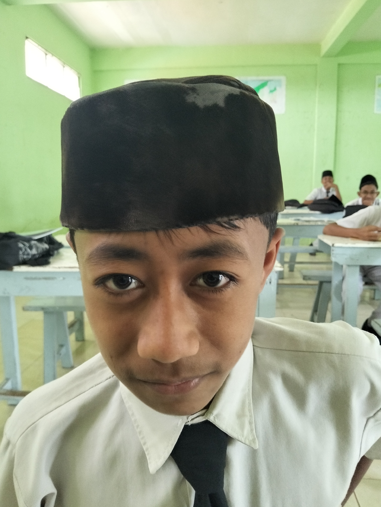 The image is a close-up portrait of a young male wearing a black songkok (a traditional cap worn by Muslim men in Southeast Asia) and a light-colored collared shirt with a dark tie. He is indoors, likely in a classroom, with light green walls and windows visible in the background. Other students and desks are also visible in the blurred background. The lighting appears to be natural, coming from the windows. The subject's gaze is directed at the camera. The overall impression is that of a candid photograph taken in an educational setting. The location of Kranji, Indonesia is not verifiable from the image itself.