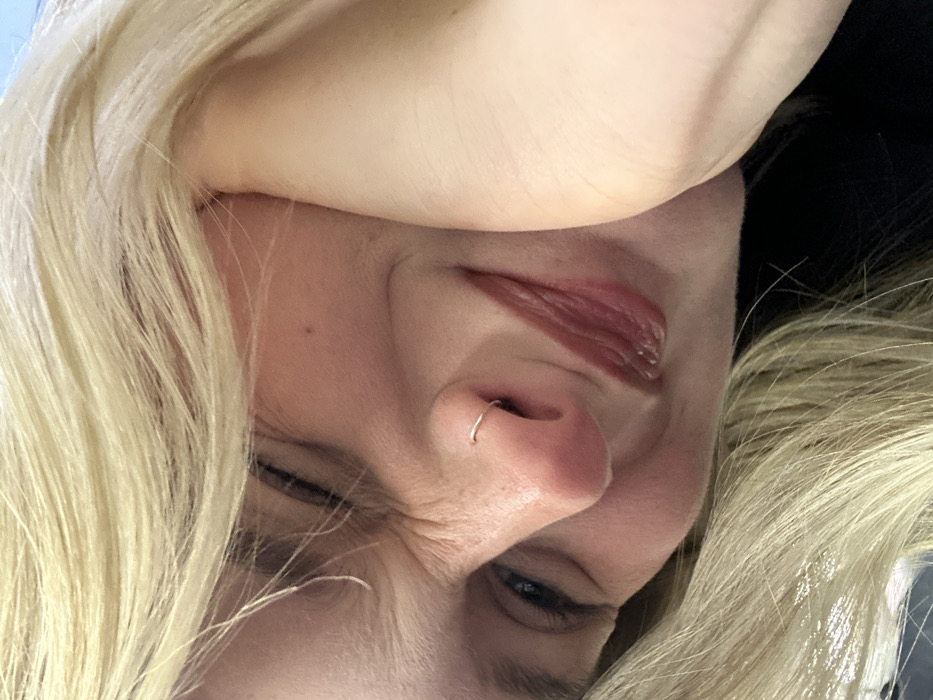 An extreme close-up shows a woman's face tilted at an angle. She has blonde hair, pale skin, and a nose ring. One eye is slightly open, showing a blue iris. The other eye is squinting. Fine lines and wrinkles are visible around the eyes. Her lips are glossy and partially open. Part of her arm or hand is visible above her head. The background is dark.