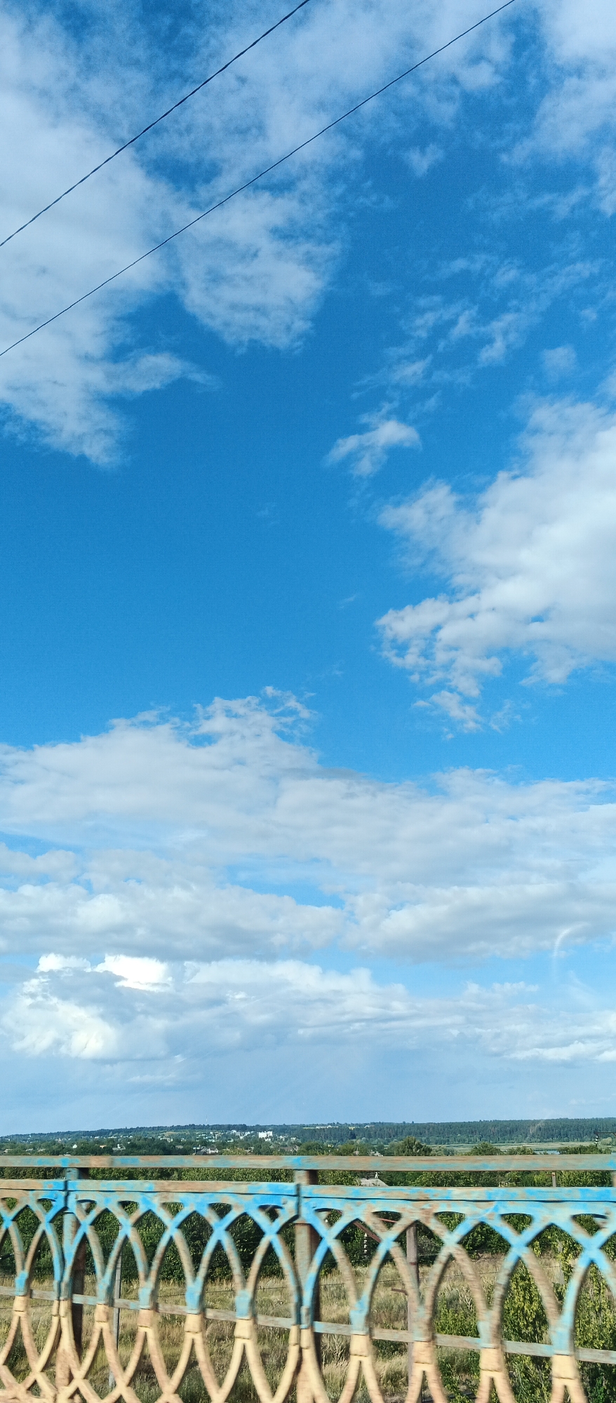 The image shows a view from a bridge over a landscape. In the foreground is the railing of the bridge, which is made of metal with a decorative pattern of arches, painted in blue and a rusty brown color. Behind the railing, a rural landscape is visible, with fields, trees, and some scattered buildings. The sky above is blue with scattered white clouds. A power line runs across the upper part of the image. The image appears to be taken in the city of Smila, Ukraine, looking out over the surrounding countryside. There are no people or animals visible in the image.