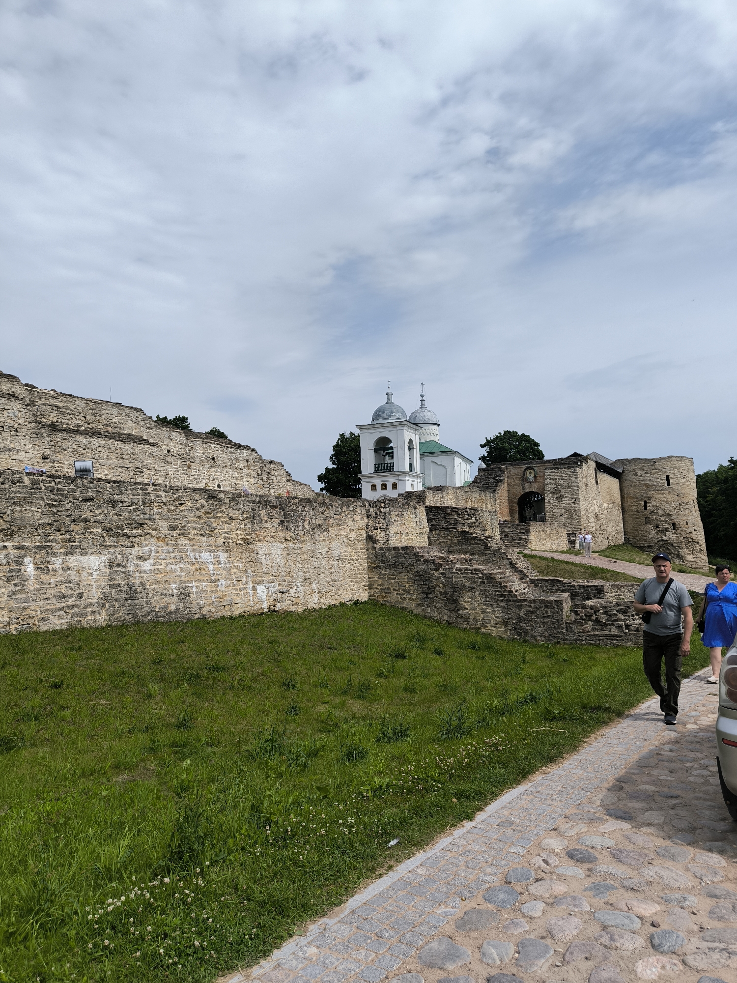 The image depicts the exterior of the Izborsk fortress in Pechory, Russia, under a partly cloudy sky. The fortress walls, constructed from rough-hewn stones, dominate the left and right sides of the frame, showing signs of age and weathering. A grassy slope with scattered white wildflowers leads up to the lower sections of the stone walls.
In the background, a white bell tower with green domes, characteristic of Russian Orthodox churches, stands prominently. Adjacent to it are stone structures that appear to be part of the fortress complex. A paved walkway, made of cobblestones, curves from the bottom right corner into the scene, leading towards an arched gateway in the fortress wall.
Two individuals are walking on the cobblestone path. In the foreground, a man wearing a grey t-shirt and dark pants is walking away from the camera, with a black bag slung over his shoulder. To his right and slightly behind him, a woman in a bright blue dress is also walking away from the camera. Their presence suggests they are tourists or visitors to the historical site. The overall scene is one of historical architecture and a visitor experience.