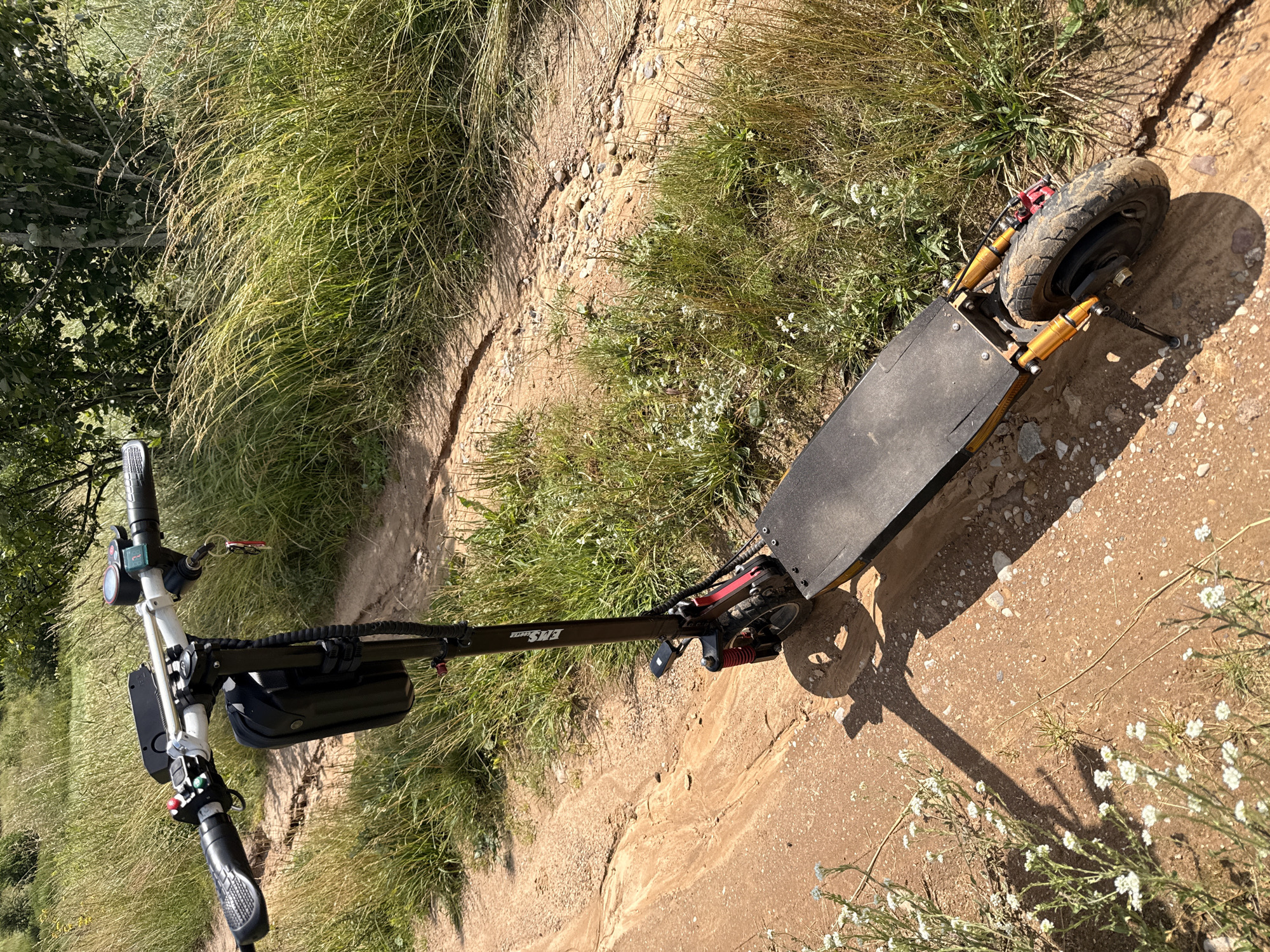 An electric scooter is parked on a dirt path in a grassy, rural area. The scooter is black and red with a black bag attached to the handlebars. The path is a light brown dirt with scattered small rocks. Tall green and yellow grass flanks the path, with some trees visible in the background. The scene appears to be outdoors on a sunny day. The scooter's brand name, "BUNSUN," is visible on the handlebar stem.