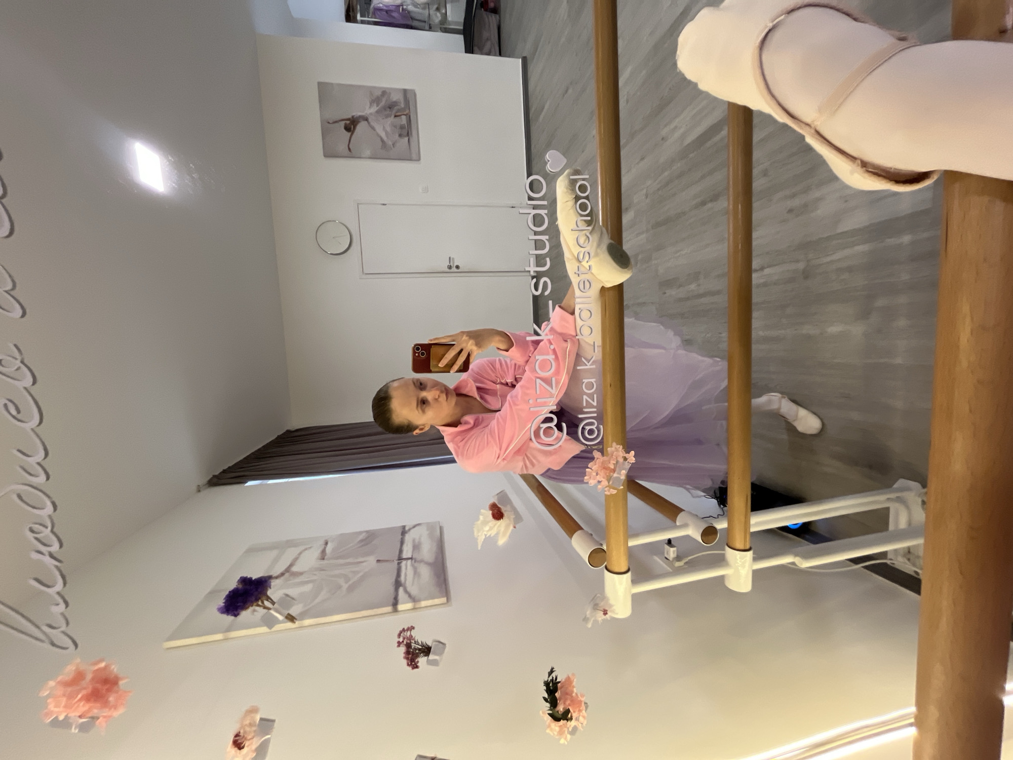 A young woman is taking a selfie in a ballet studio. She is wearing a pink pajama shirt and a light purple tutu. Her right leg is extended in a ballet position and is resting on a barre. She is holding her phone in her left hand, taking a picture of herself in the mirror. The studio has a white wall with artwork, including ballet-themed images and floral decorations. In the background, there is a door and a window with blinds. The location is likely in a ballet studio, potentially in Budva, Montenegro.