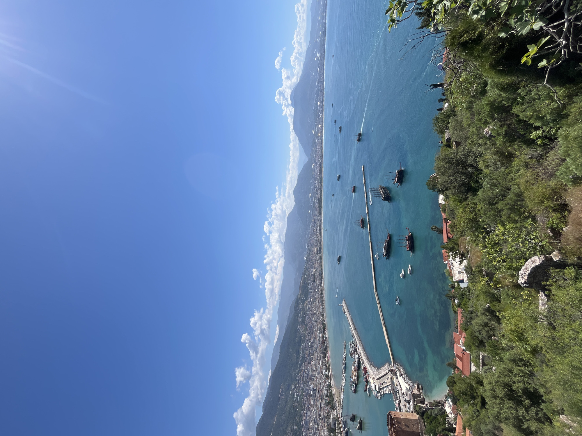 The image captures a panoramic view of Alanya, Turkey, from a high vantage point. The scene is dominated by the turquoise sea, where several boats are scattered. A long pier stretches into the water, leading to a bustling harbor filled with yachts and smaller vessels. Along the coastline, buildings and houses are visible, nestled between the mountains and the sea. The sky is a clear blue with fluffy white clouds on the horizon. Lush green vegetation covers the foreground, suggesting the location is a high point, such as a castle or a hill overlooking the city.